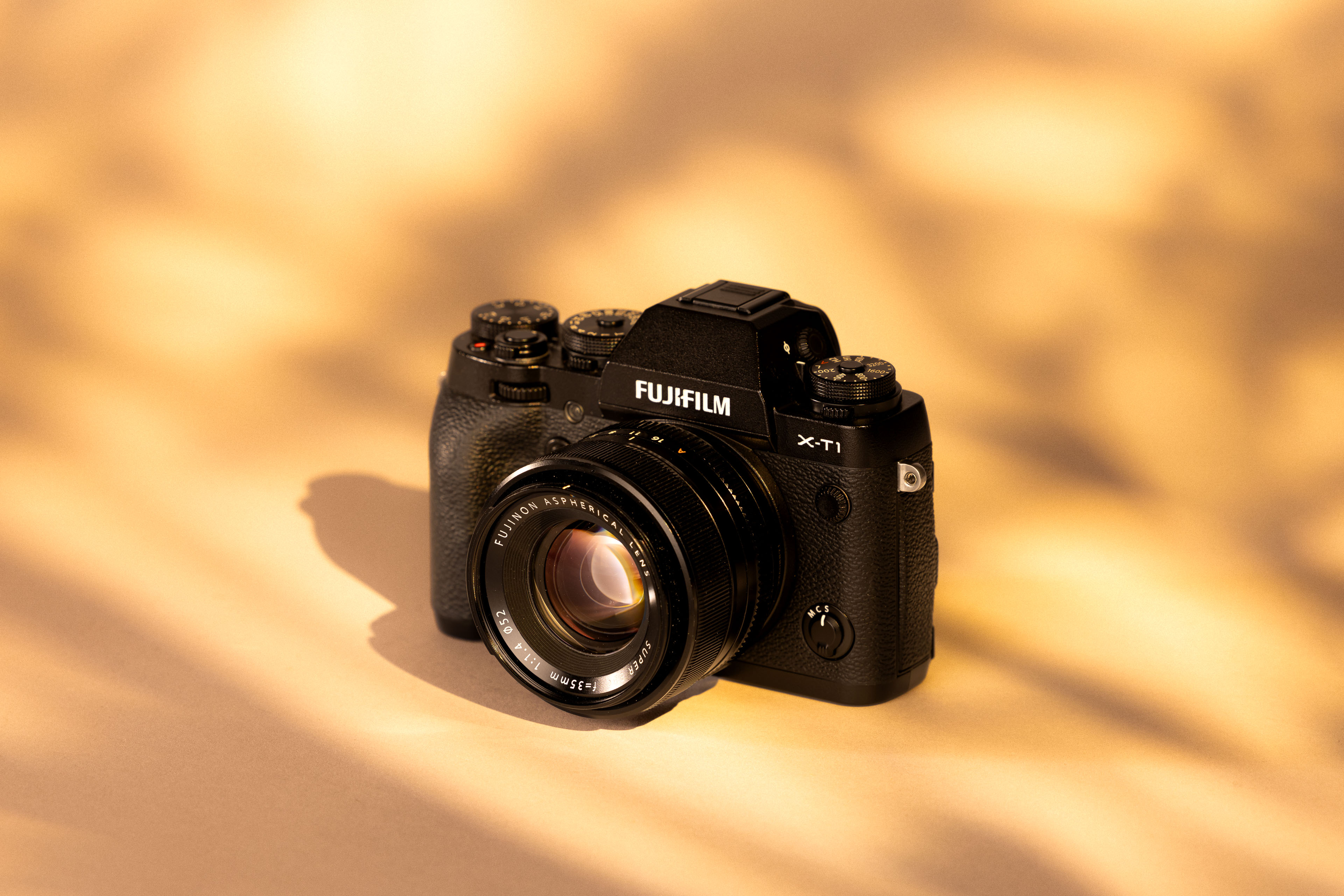 A Fujifilm X-T1 with a Fujifilm XF 35mm f/1.4 R lens. The camera is photographed on a sun-dappled background.