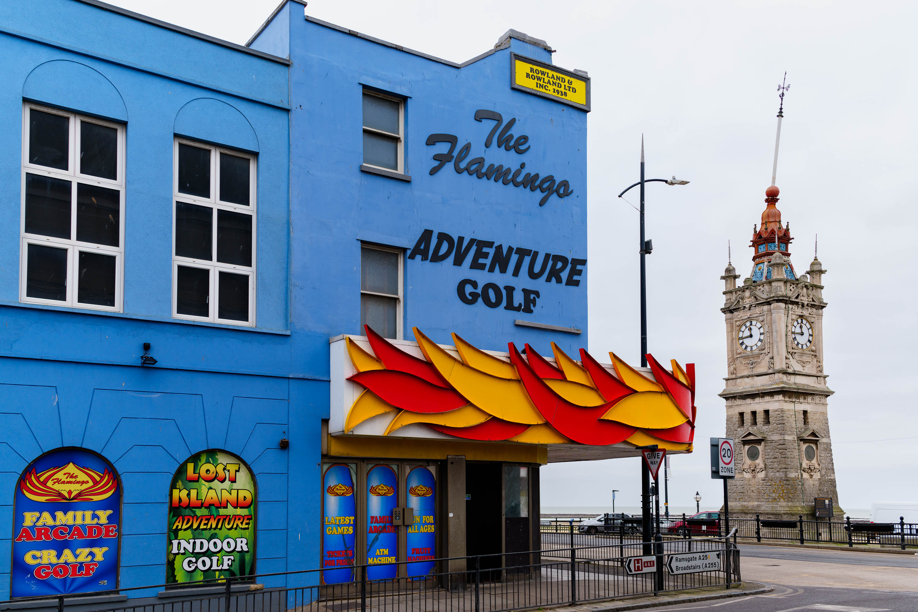 Ein leuchtend blaues Gebäude an einem bedeckten Tag am Meer, das mit farbenfroher Beschilderung und einem Vordach mit Flammendekor für „The Flamingo Adventure Golf“ wirbt. Im Hintergrund, ein historischer Uhrenturm. Aufgenommen von Connor Redmond mit einer Sony RX1R III.
