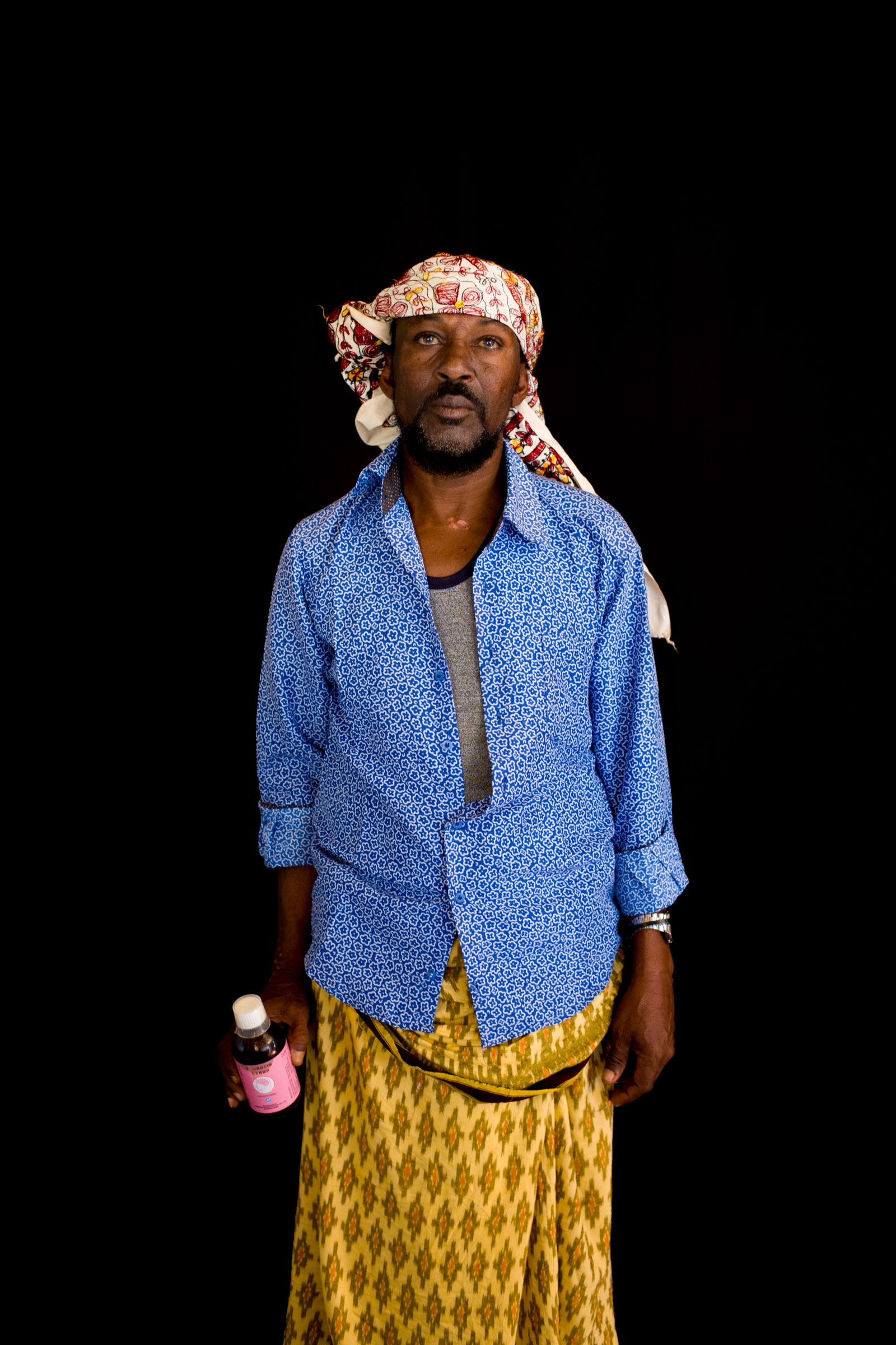 Un homme vêtu de vêtements lumineux tenant une bouteille en plastique regarde directement vers l'appareil photo, se tenant contre un fond sombre.