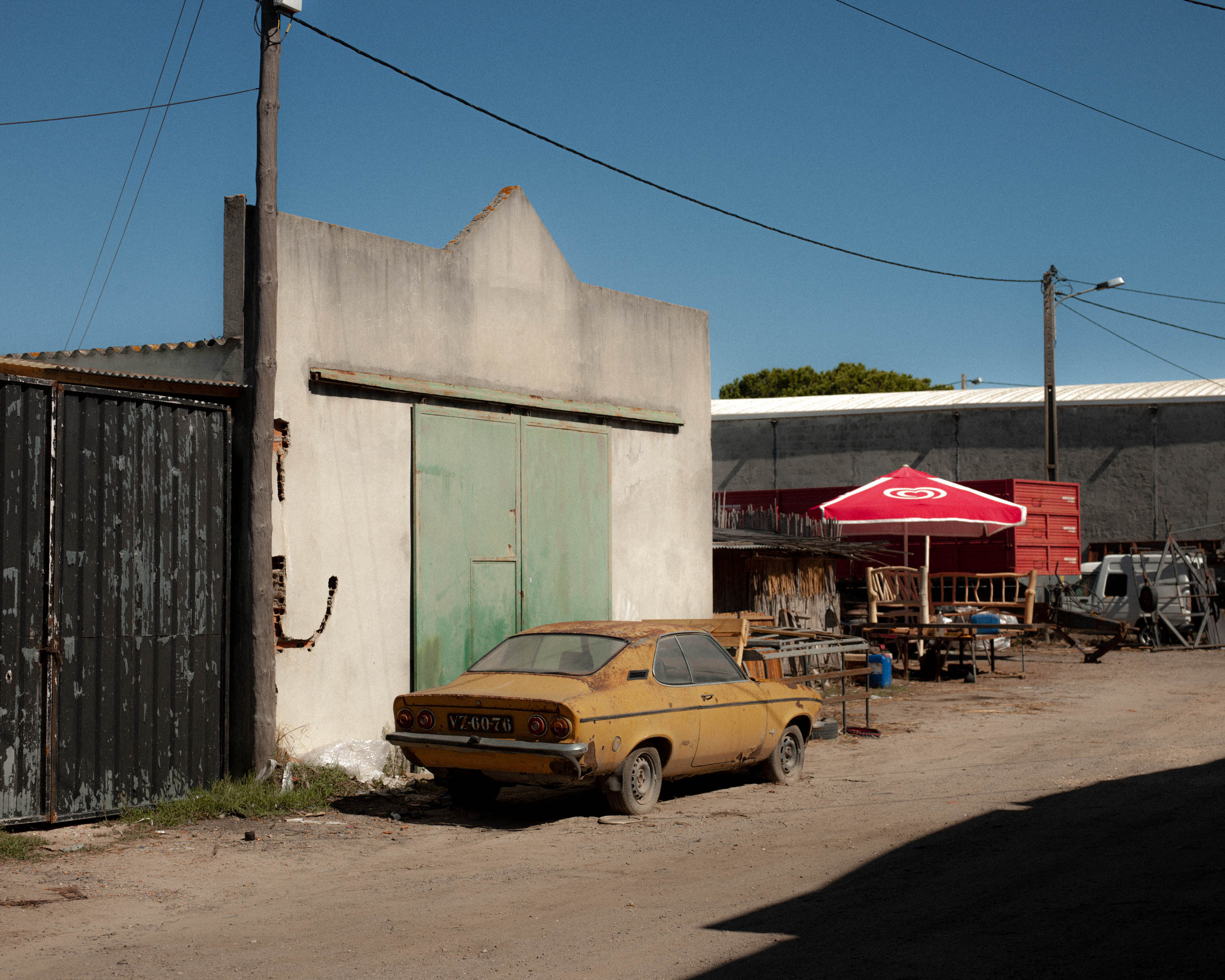 A yellow car parked on a dusty road with a blue sky and red parasol in the distance, shot on Phase One P45 Plus by Ian Howorth