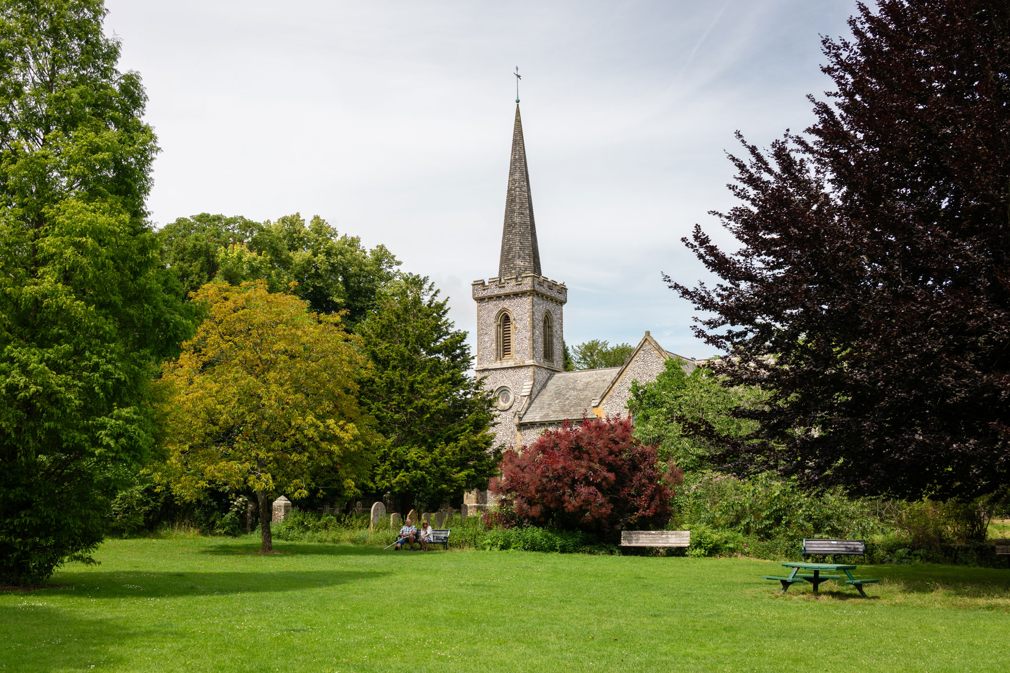 A church with a steeple can be seen behind a park's greenery, with two people sitting on one of four benches. Light wispy clouds cover a blue sky. 