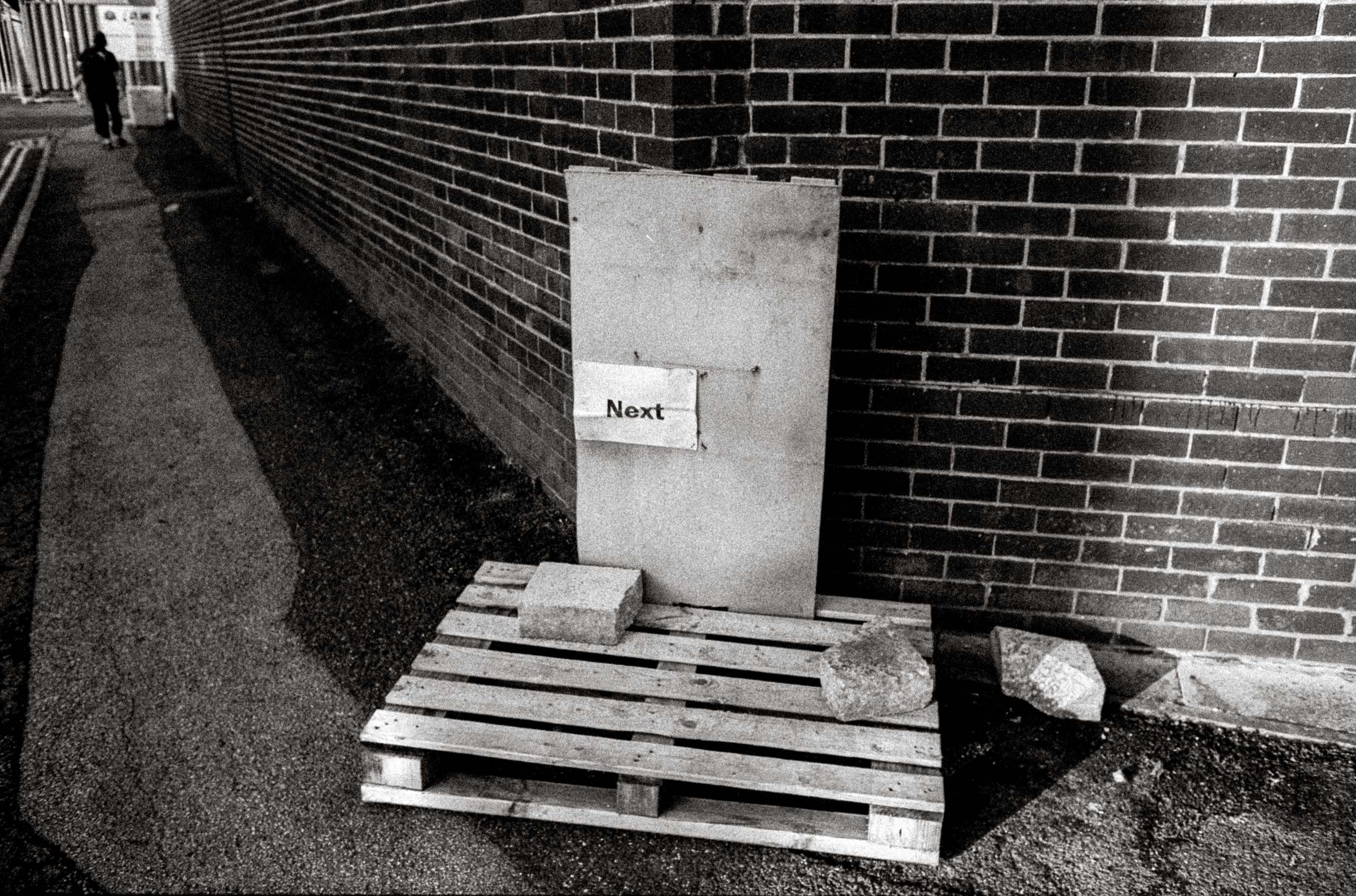 A pallet in front of a brick wall with a piece of paper with the word ‘Next’ printed on it, black-and-white street photography by Ian Howorth