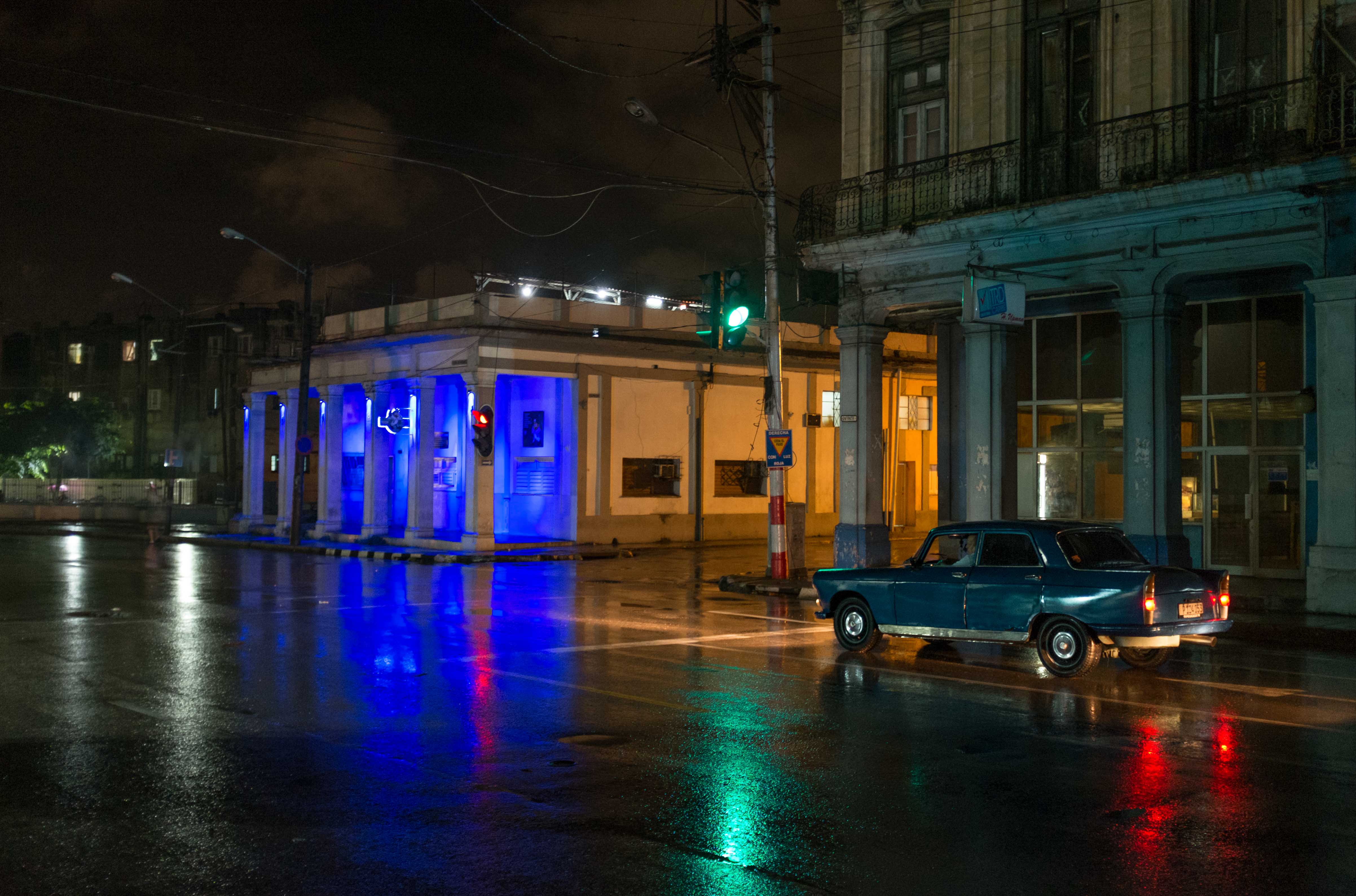 Photo of an old dark-green car driving at night as white, blue, green and red lights reflect off the wet road, street photography by Ian Howorth