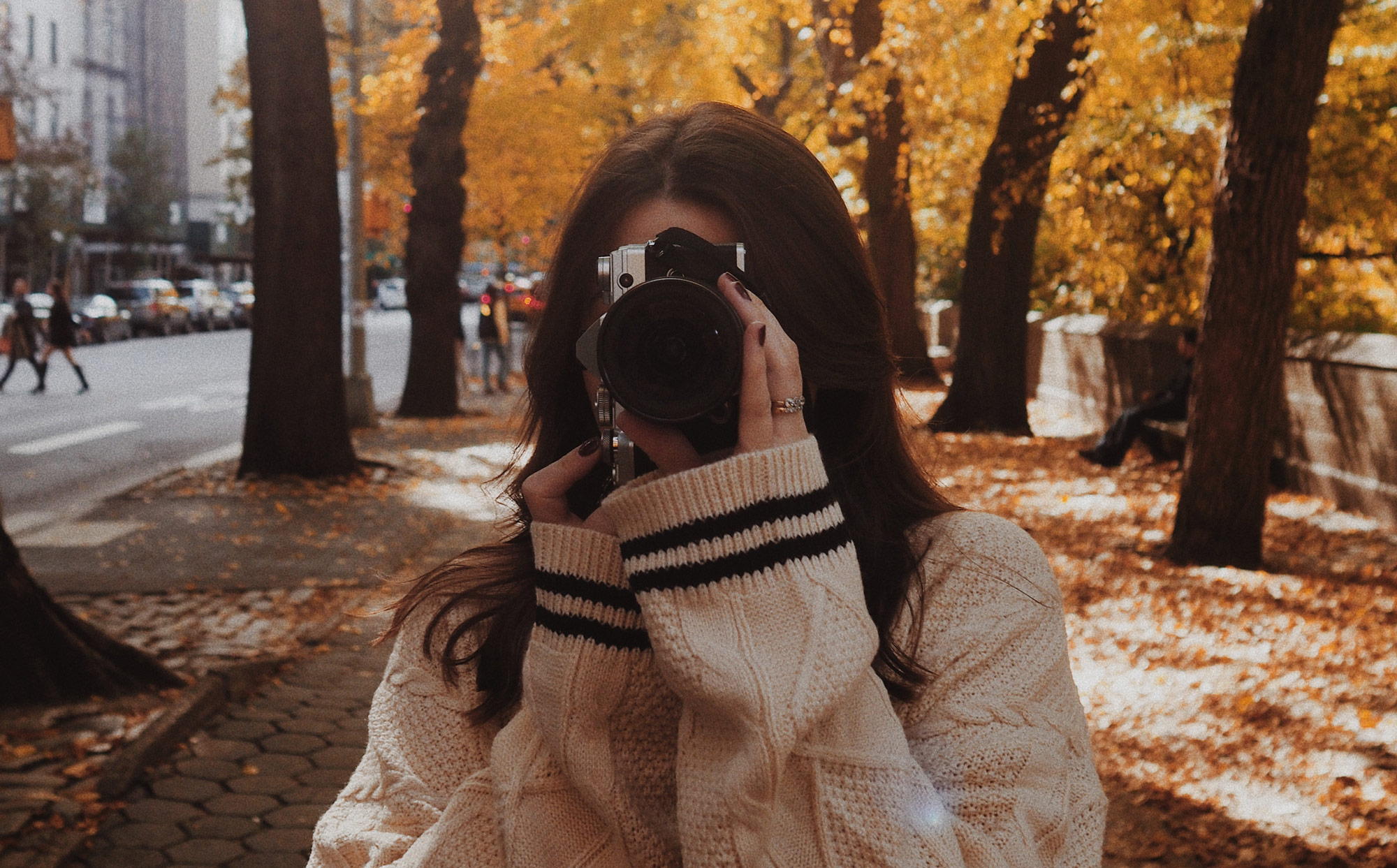 A cropped photo of Georgina Lamrock taking a photo with the Olympus E-M5 III  in Central Park with an avenue of trees and a road in the background 