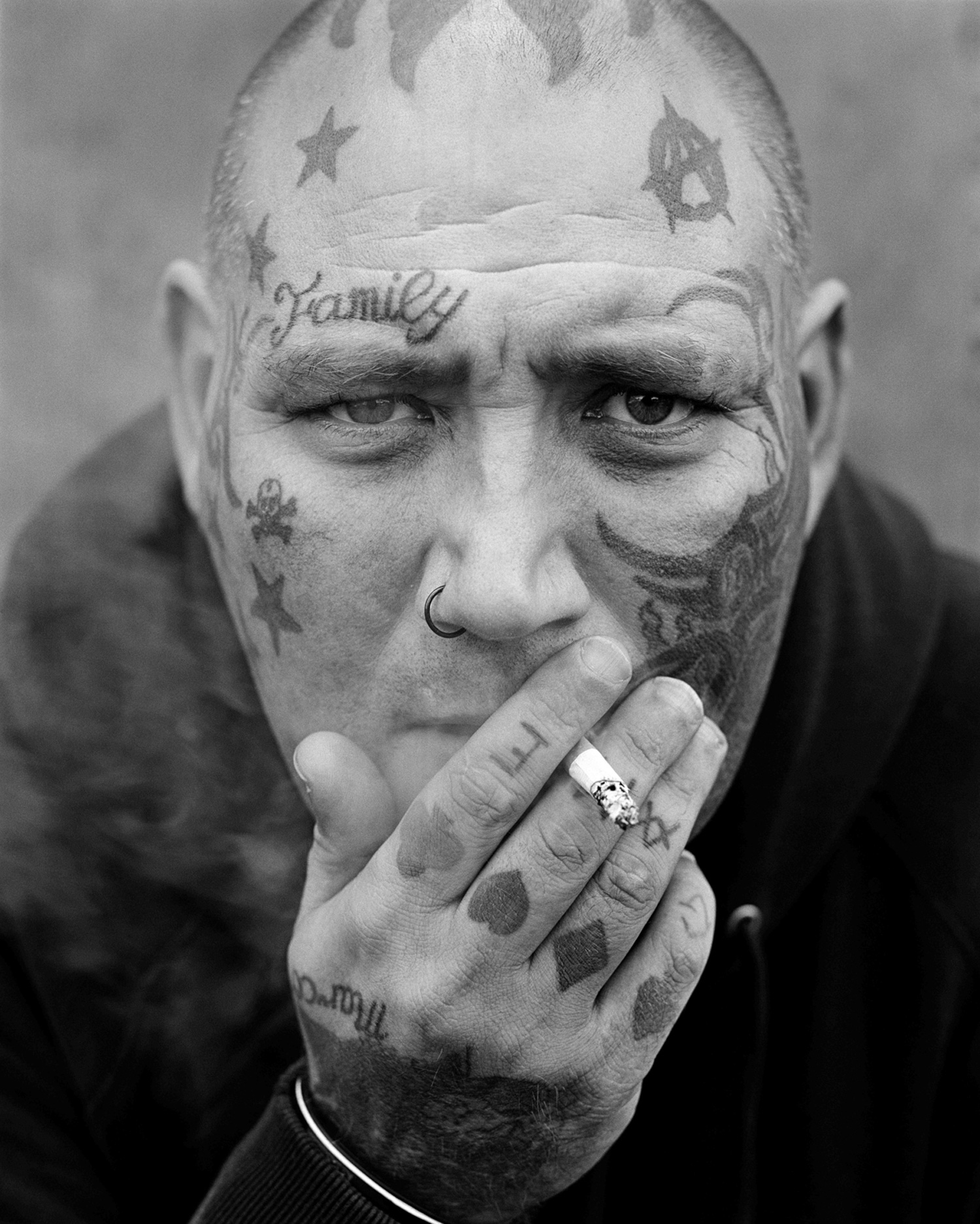 Black-and-white close-up street portrait of heavily tattooed person smoking, image by Simon Murphy