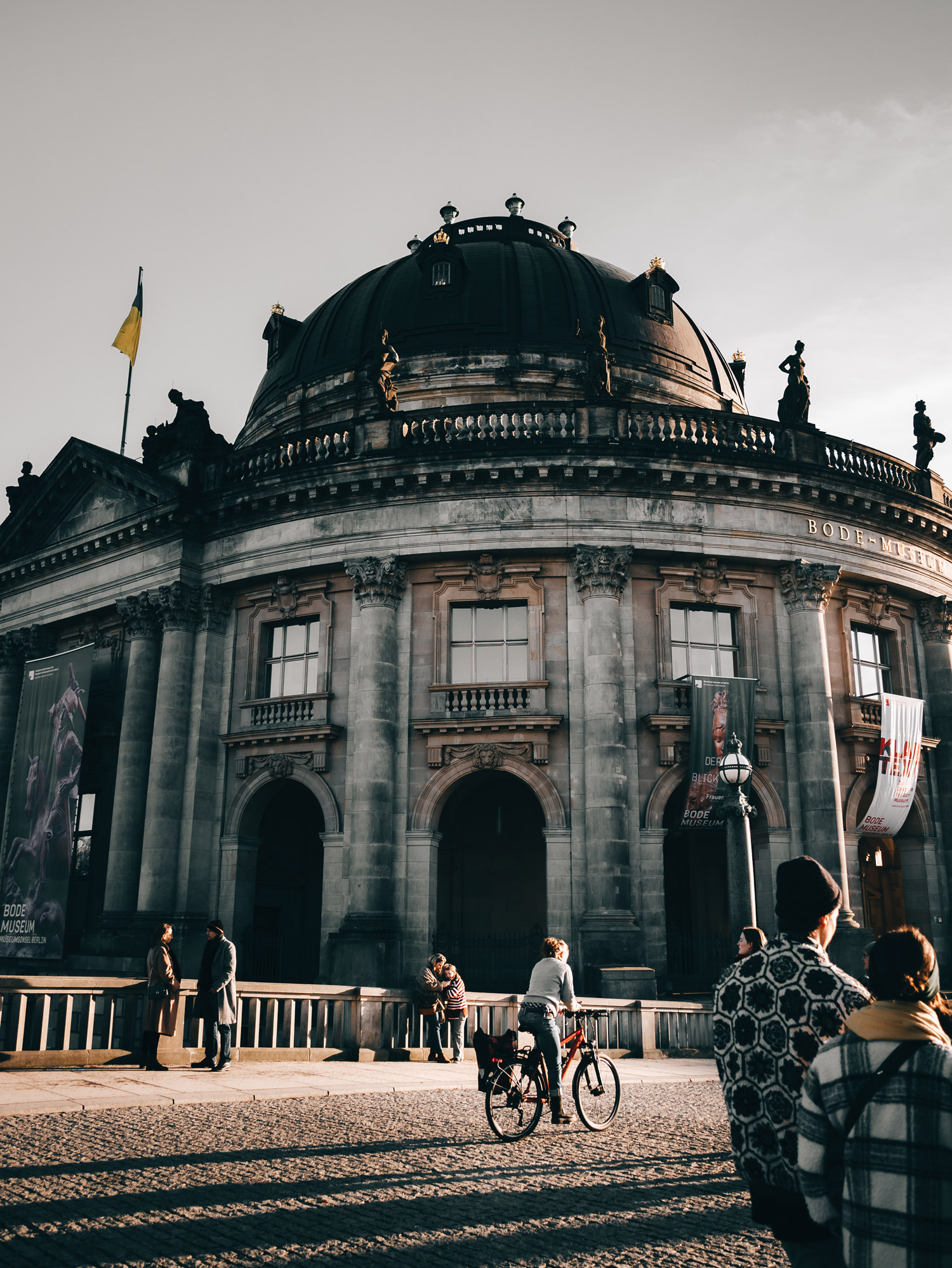 Ein altes Steingebäude mit einer Person auf einem Fahrrad und Fußgänger:innen, die daran vorbeifahren, fotografiert von Emily Lowrey mit der Panasonic Lumix DC-GX800.