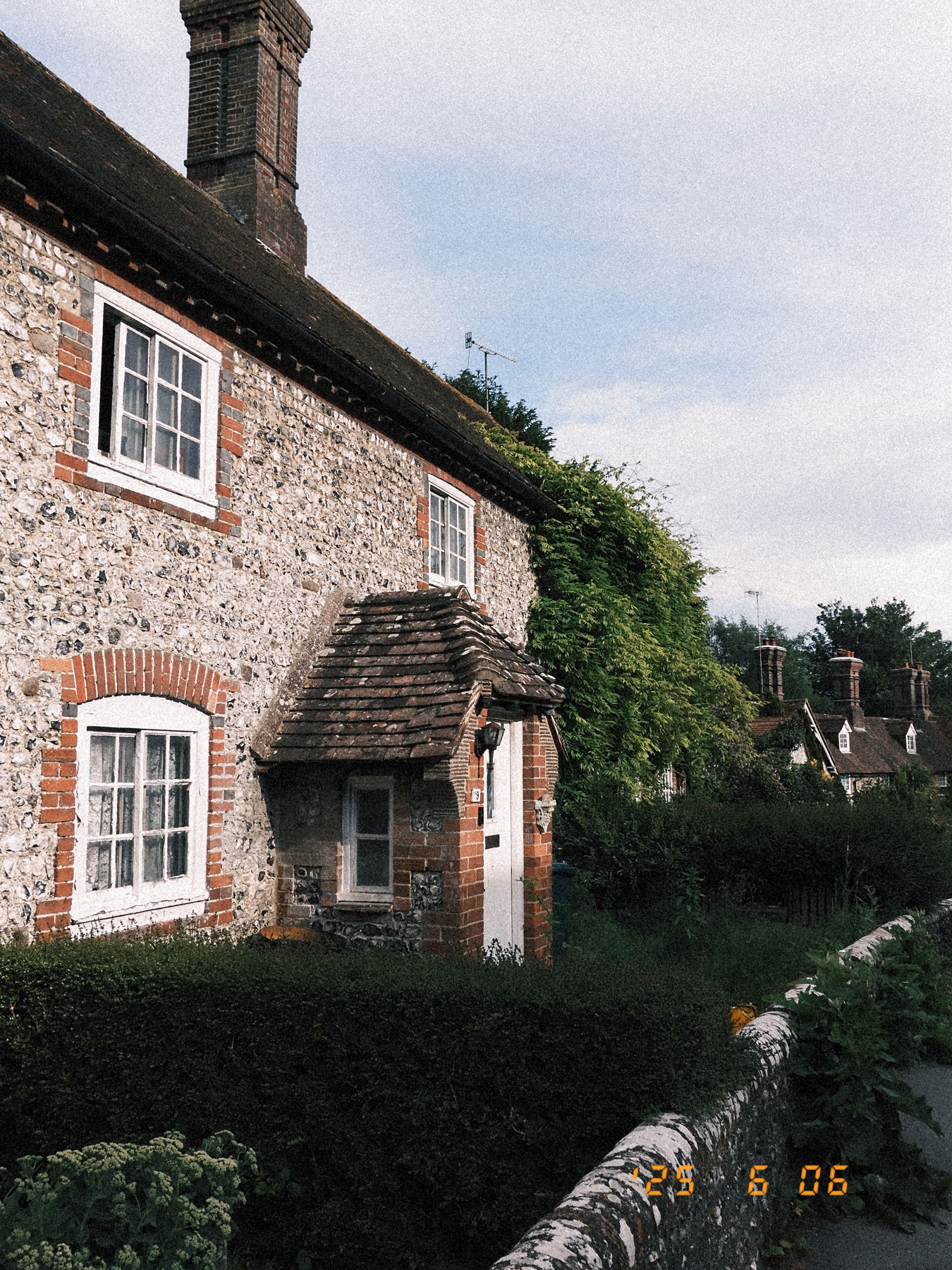 Von der Seite aufgenommene Fassade eines alten Hauses mit weiß gerahmten Fenstern. Im Hintergrund, eine Häuserreihe. Im Vordergrund, eine Gartenmauer und ein Vorgarten.