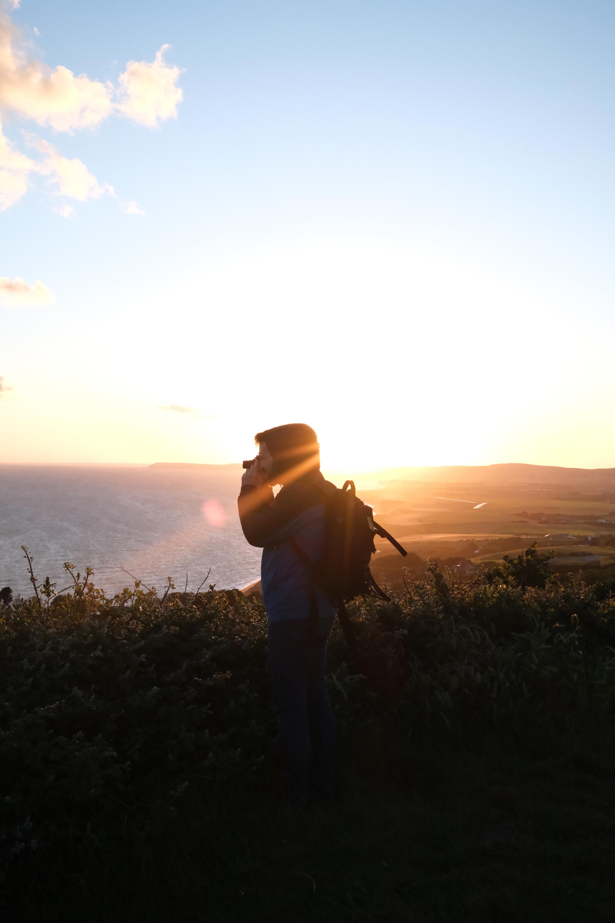 MPB videographer Jakub Golis takes some photos surrounded by greenery on a hillside on the Isle of Wight. Behind him, the sun is setting and the sea is calm.