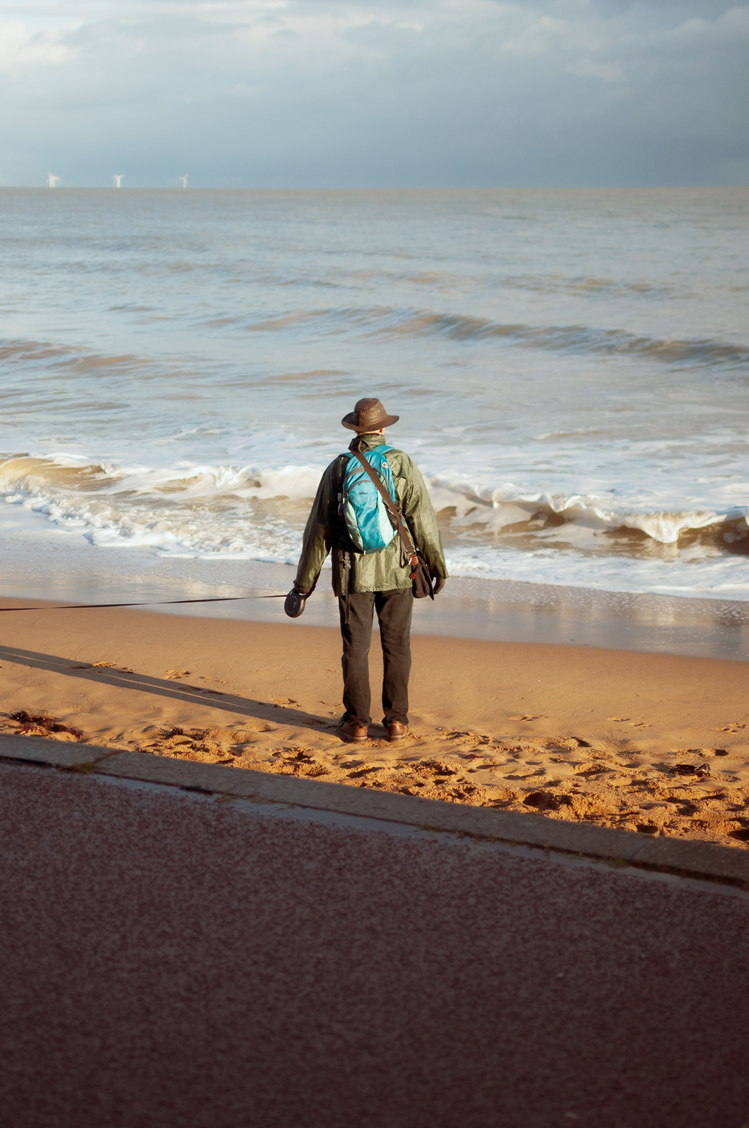 A person looking out to sea holding a dog lead and wearing a wide-brimmed hat. By Connor Redmond.