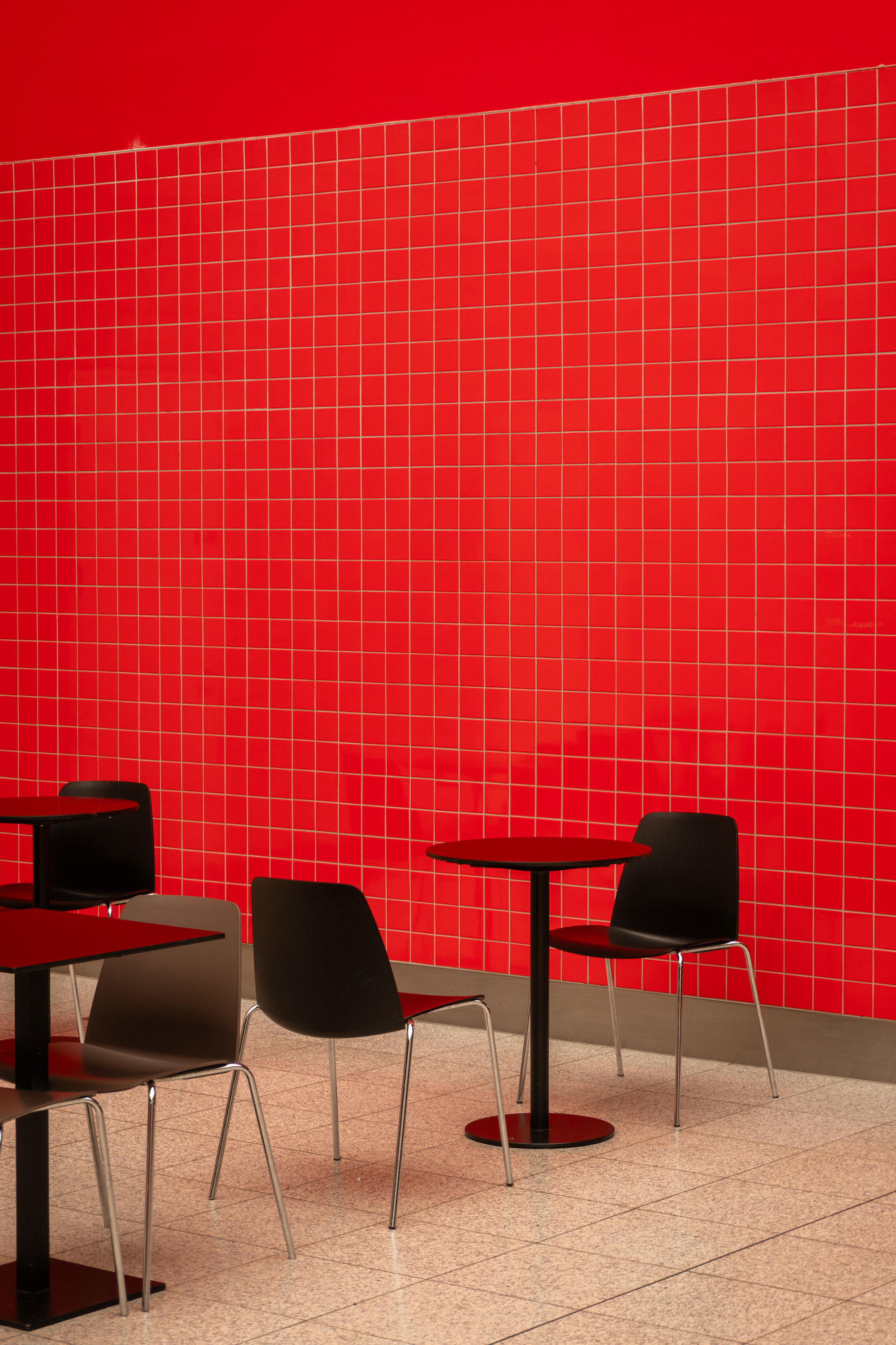 Zwei leere, mattschwarze Stühle und ein Tisch auf gefliestem Boden. Im Hintergrund eine rot geflieste Wand.