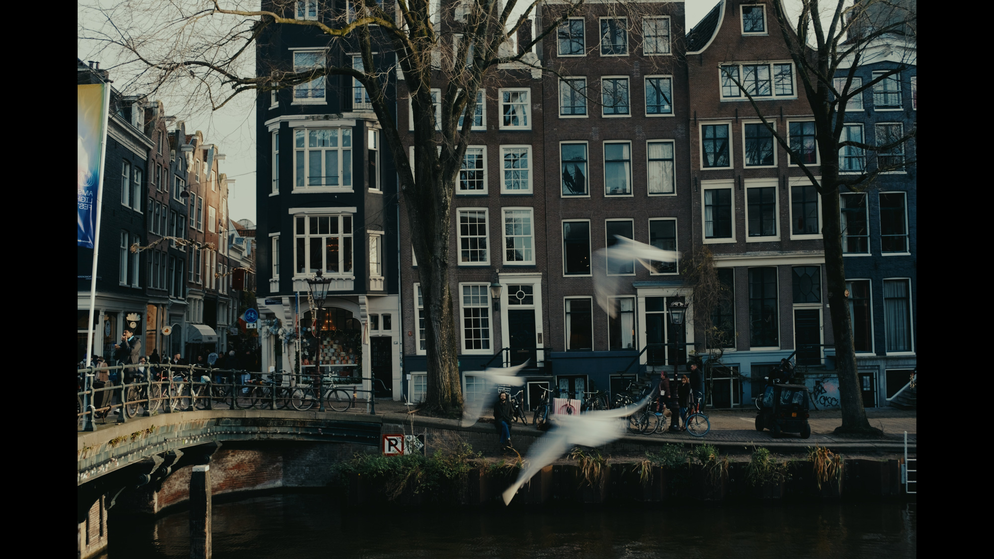 Several historic canal houses stand alongside a canal in Amsterdam, featuring classic architecture, large windows, and a bridge with parked bicycles.