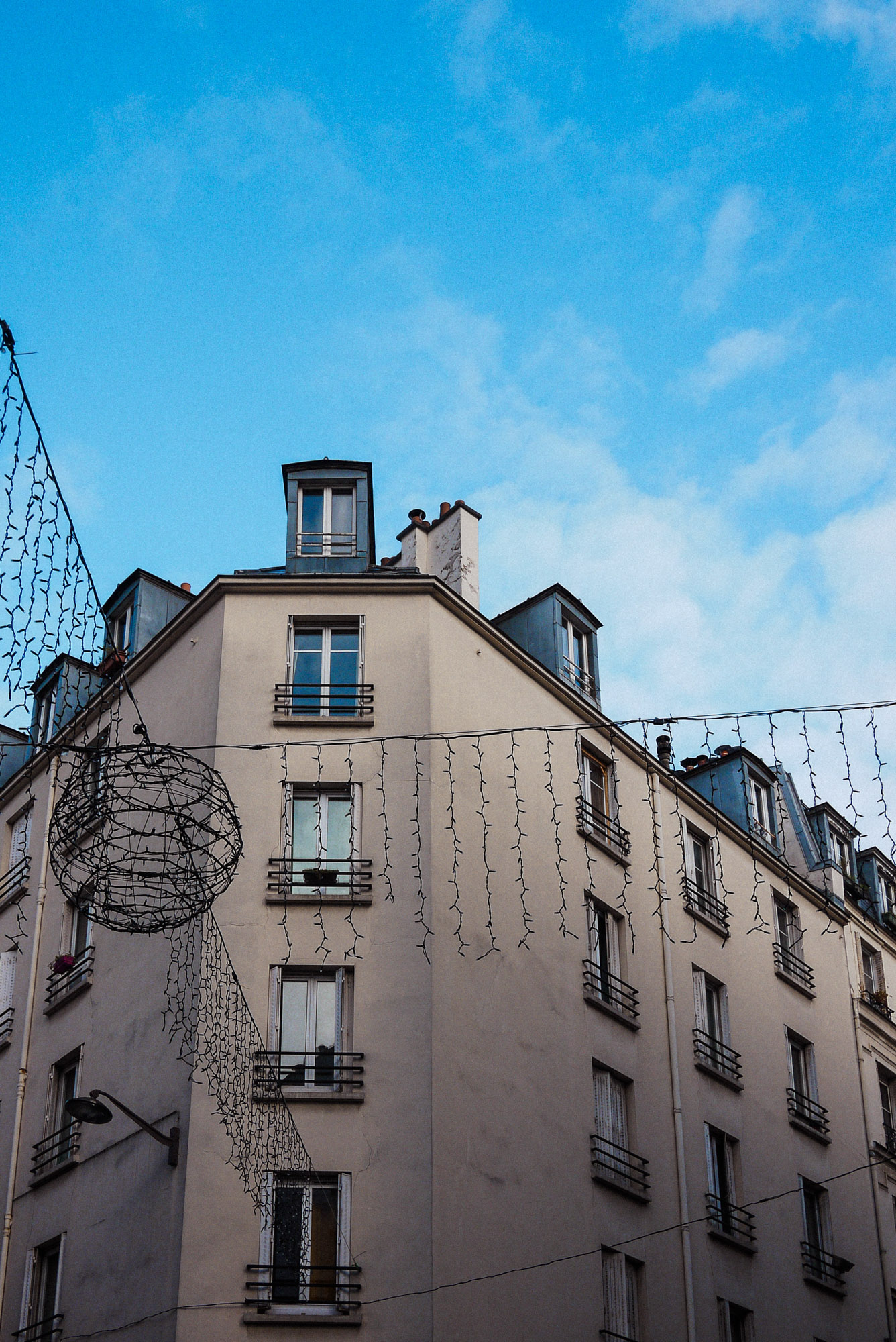 Streetaufnahme, die festliche Straßenbeleuchtung vor einem Pariser Wohnhaus mit mehreren Fenstern unter strahlend blauem Himmel zeigt. Aufgenommen von Wesley Verhoeve mit einer Panasonic Lumix GF1.