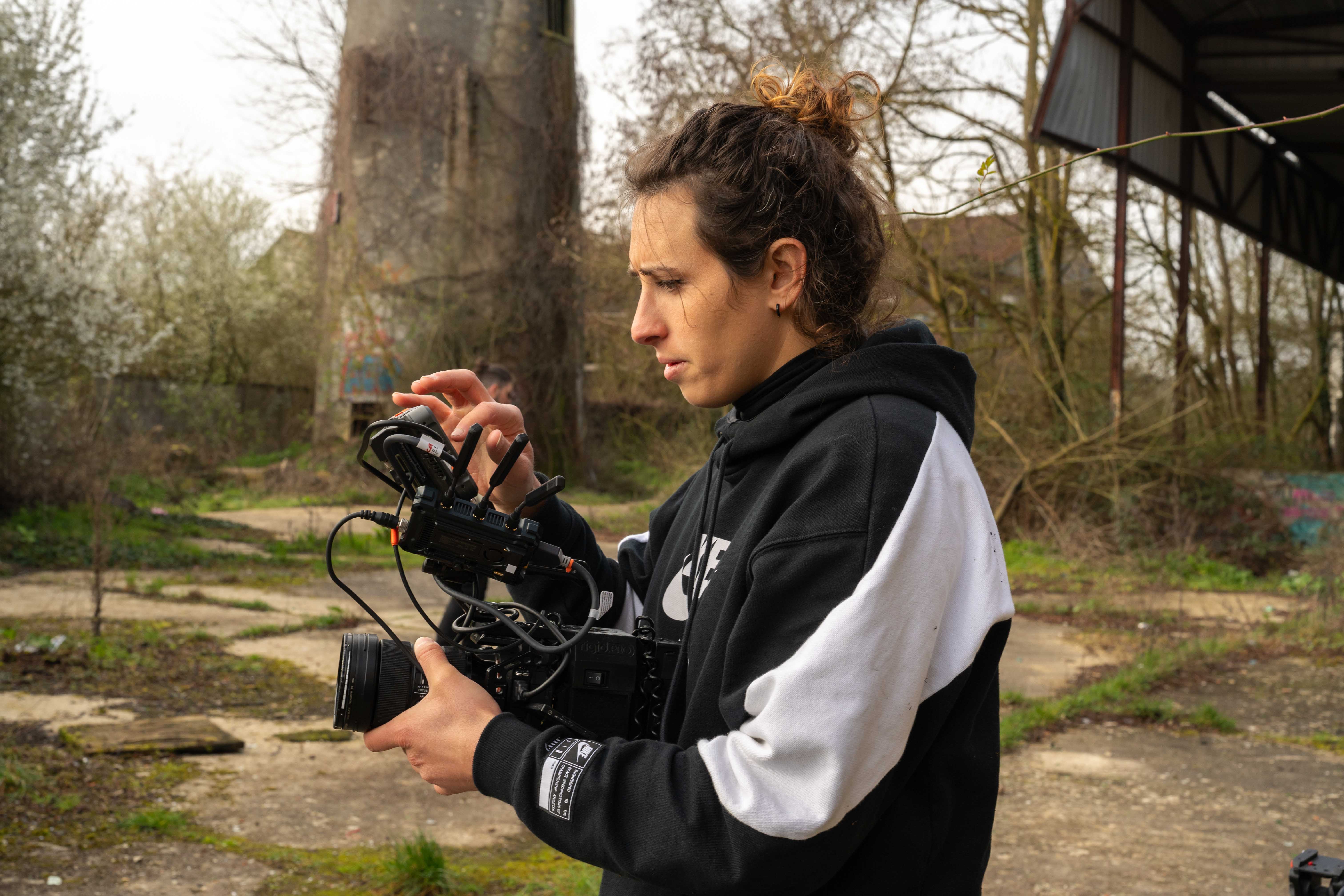 Videographer Lena Hempel adjusts a professional camera rig while standing outdoors at an overgrown industrial location.