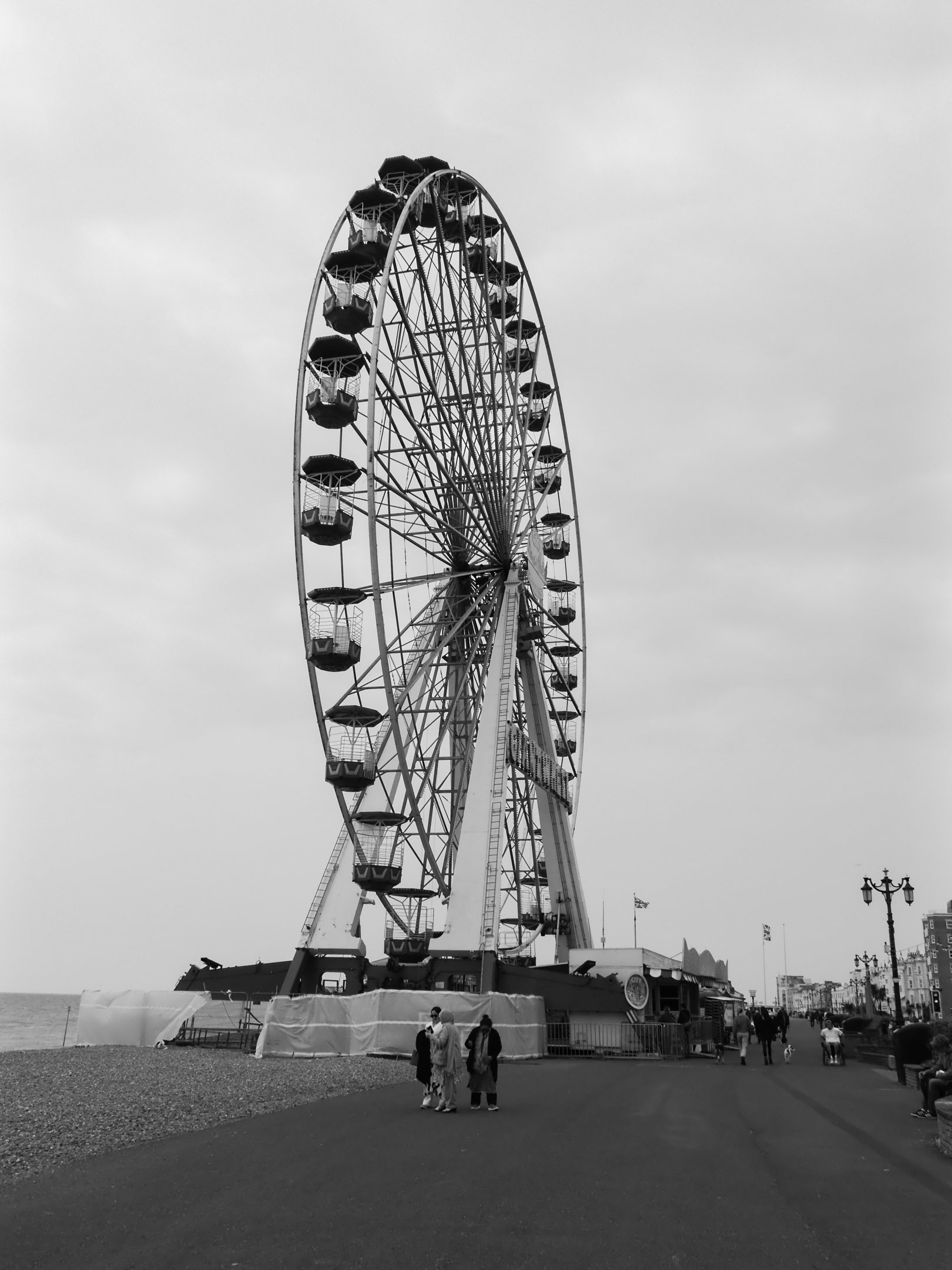 Schwarz-Weiß-Aufnahme eines Riesenrads an der Strandpromenade von Worthing. Eine Gruppe von Menschen unterhält sich im Vordergrund. Im Hintergrund, mehrere Personen.