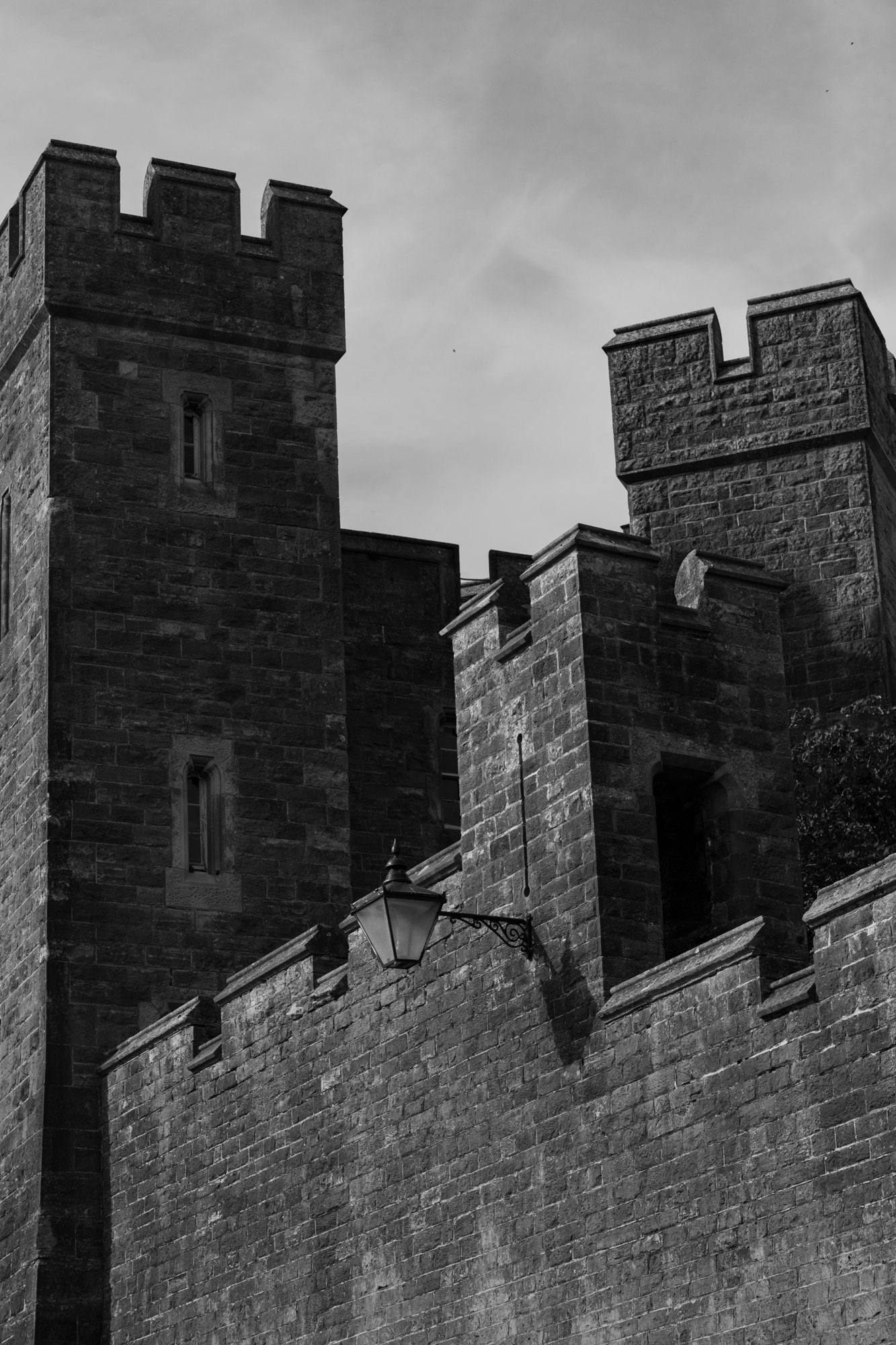 A black and white photo of a castle’s turrets. A lamp is on the side of the wall, and wispy grey clouds can be seen in the sky above.