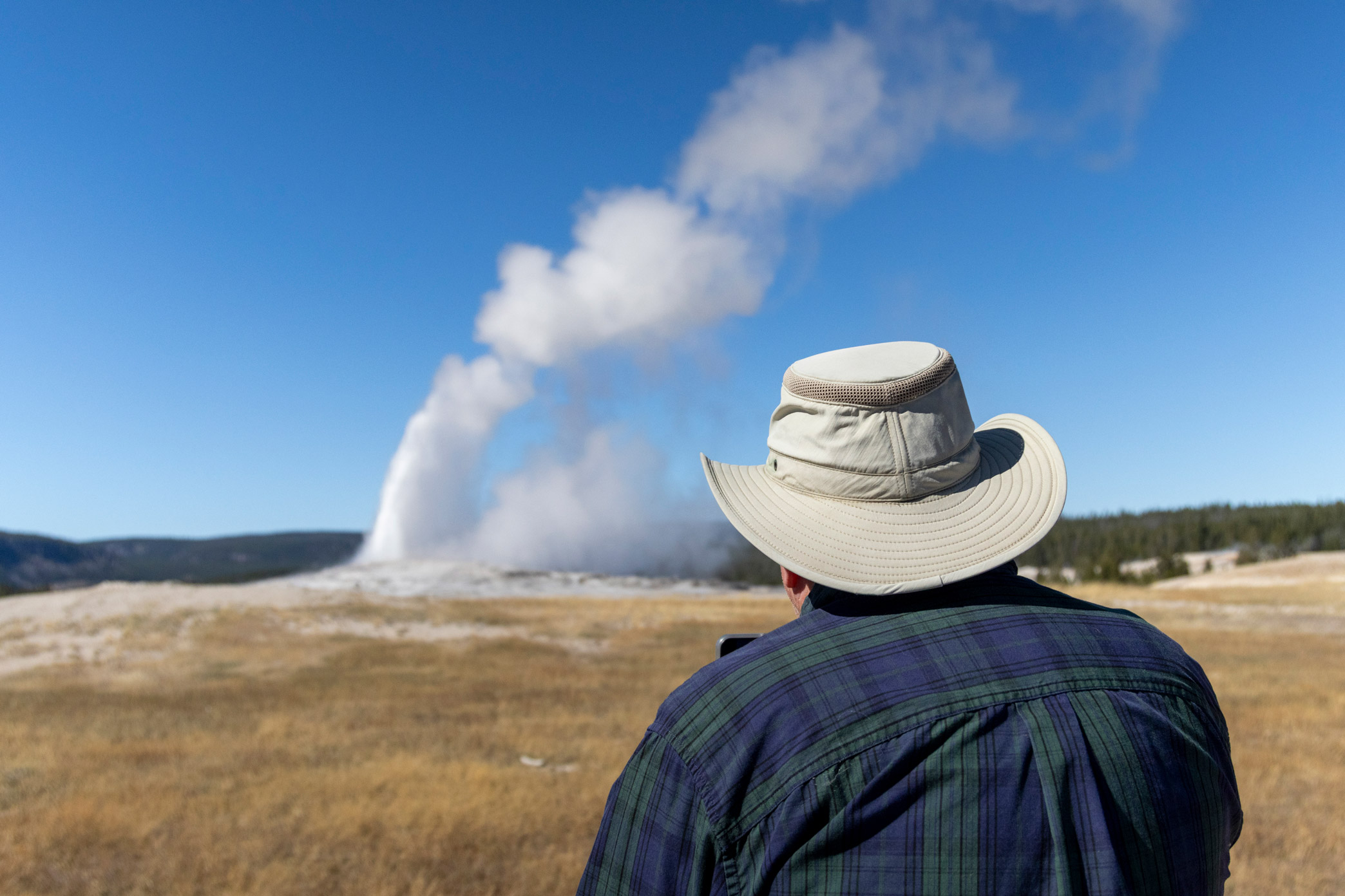 Von hinten aufgenommene Person mit Hut, die den ausbrechenden Old Faithful Geysir im Yellowstone National Park fotografiert. Aufgenommen von Kristi Townsend mit einer Canon EOS R7.