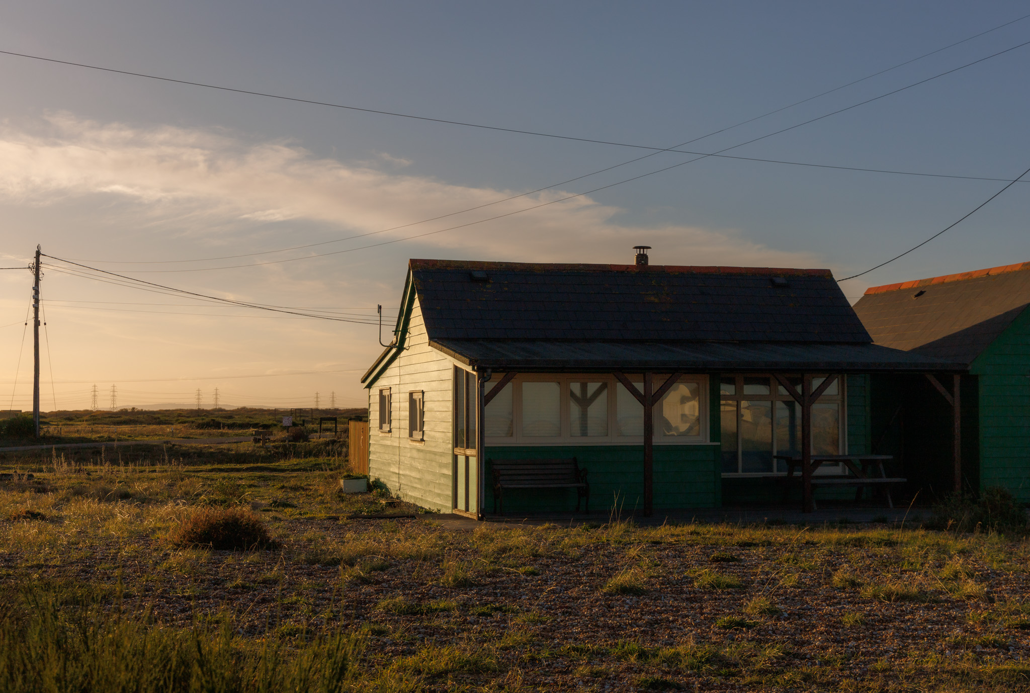 Ein kleines Haus in Dungeness bei Sonnenuntergang mit Telefonmasten und Stromleitungen im Hintergrund. Aufgenommen von Connor Redmond.