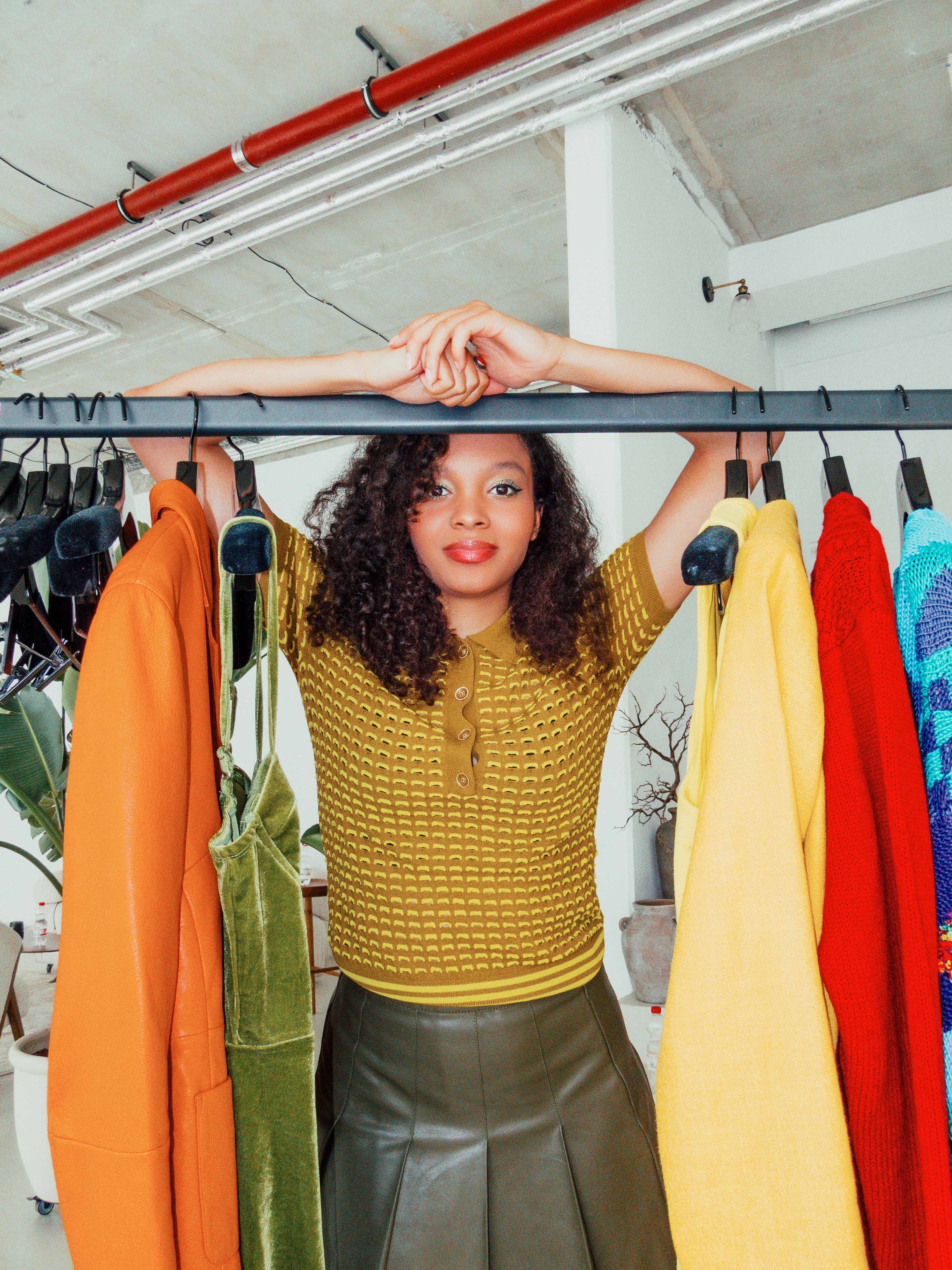 A model posing behind a rack of vibrant clothes, including orange, green, yellow, and red pieces. Shot by Alina Rudya on a Nikon Coolpix A.