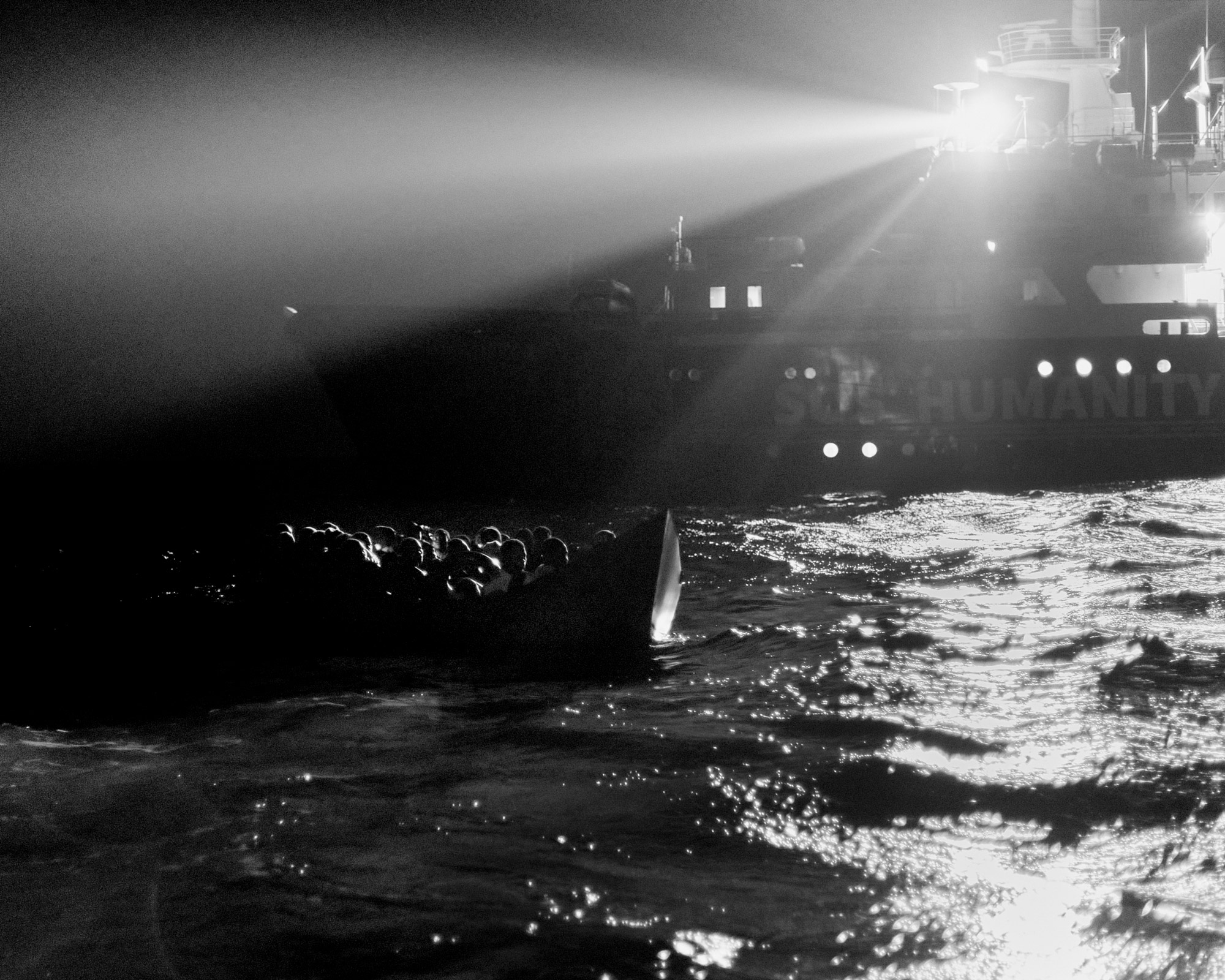 Black and white shot of a small boat at sea, in the background a large rescue ship using a high-powered spotlight.