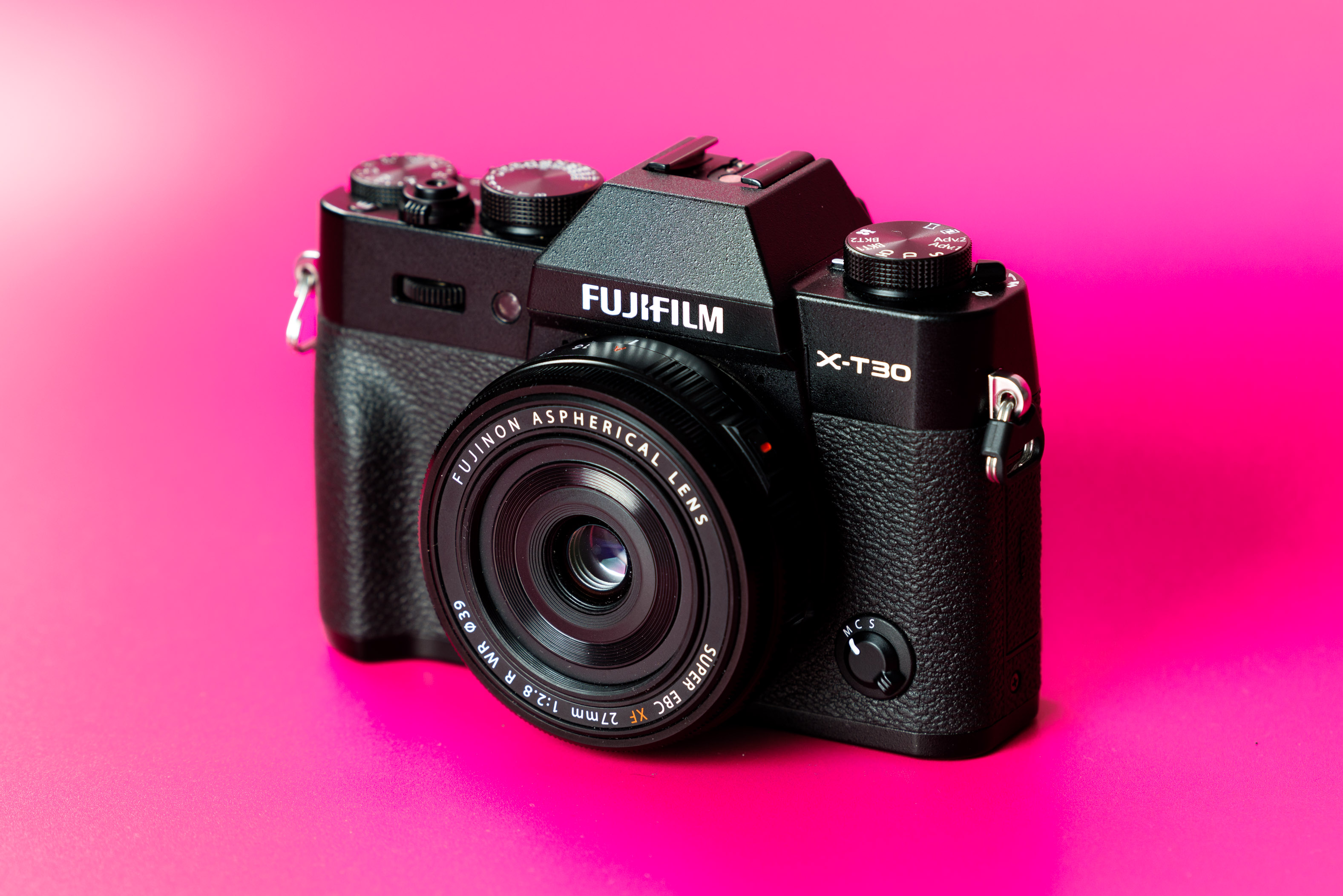 Close-up of a Fujifilm X-T30 mirrorless camera with Fujinon lens on a vibrant pink background, highlighting design, controls and compact photography gear aesthetics.