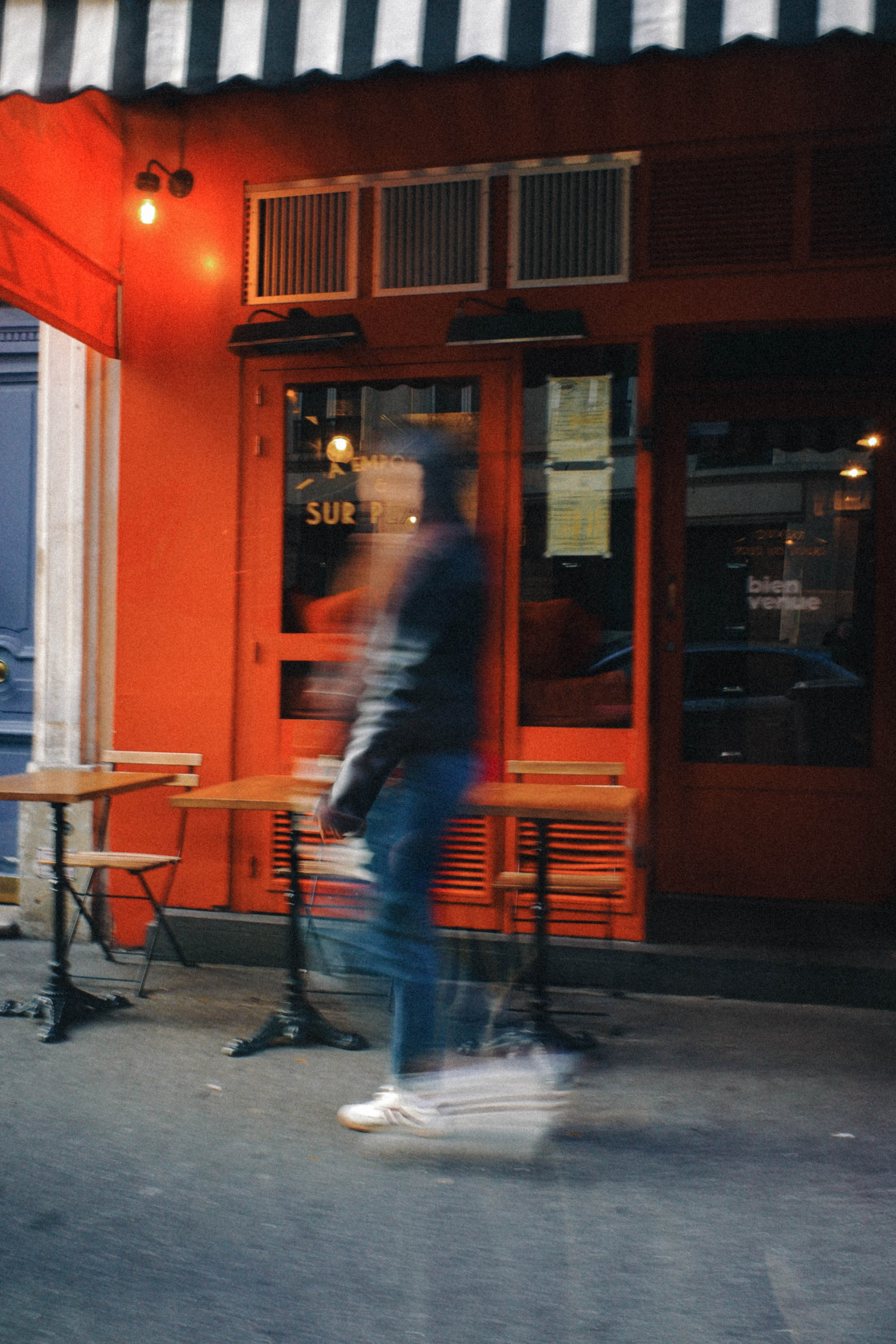 A blurred passer-by walks past a bright red Paris café, creating a cinematic street scene full of colour, movement and classic urban charm. Shot by Wesley Verhoeve on a Fujifilm XF10.