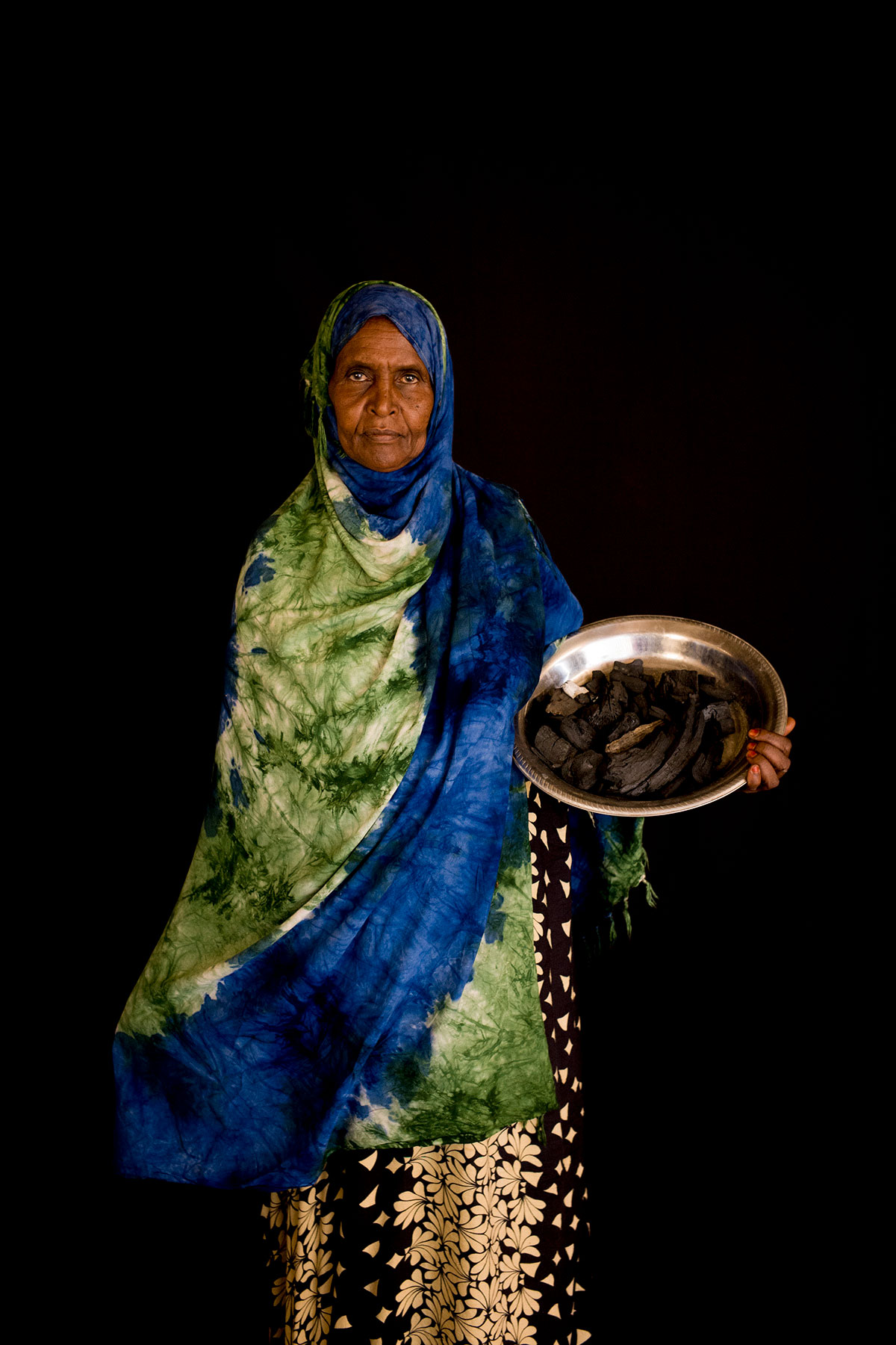 A person in bright clothes holding a large silver plate of food looks directly at the camera and stands against a dark backdrop.