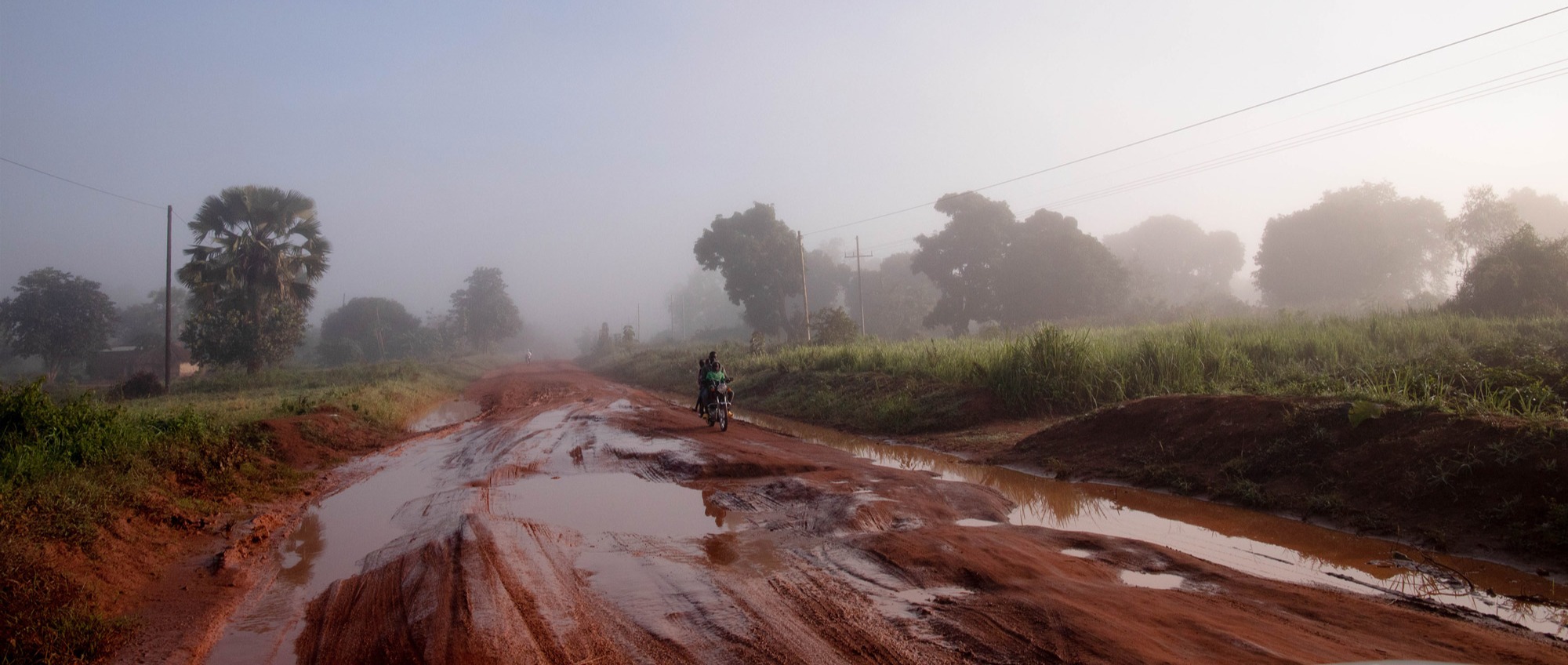 A muddy road in Uganda, with trees in the background and a moped being ridden down the road.

