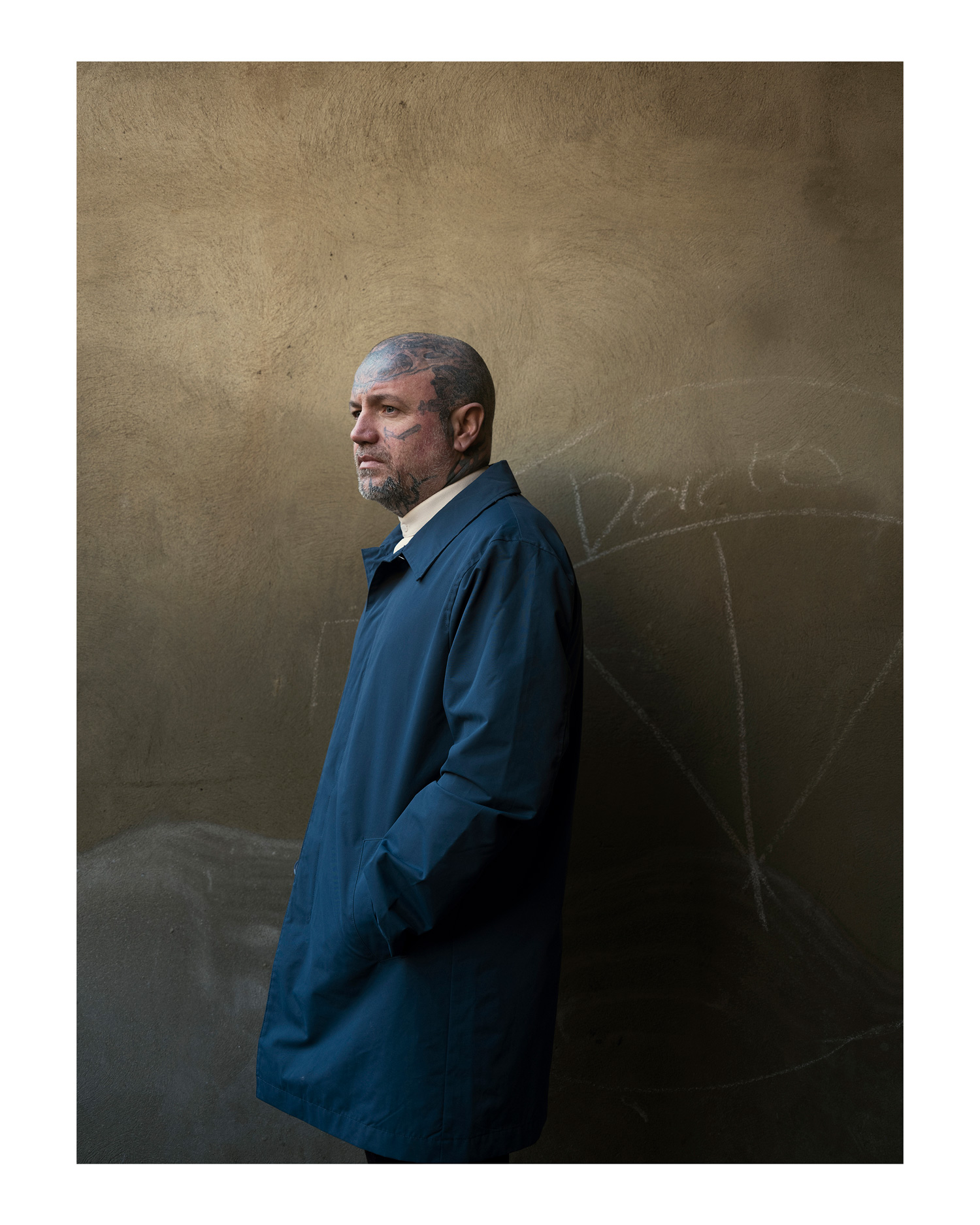 An upper-body portrait of a tattooed model wearing a blue coat standing in front of a wall, photo by Simon Murphy with the Fujifilm GFX 50S