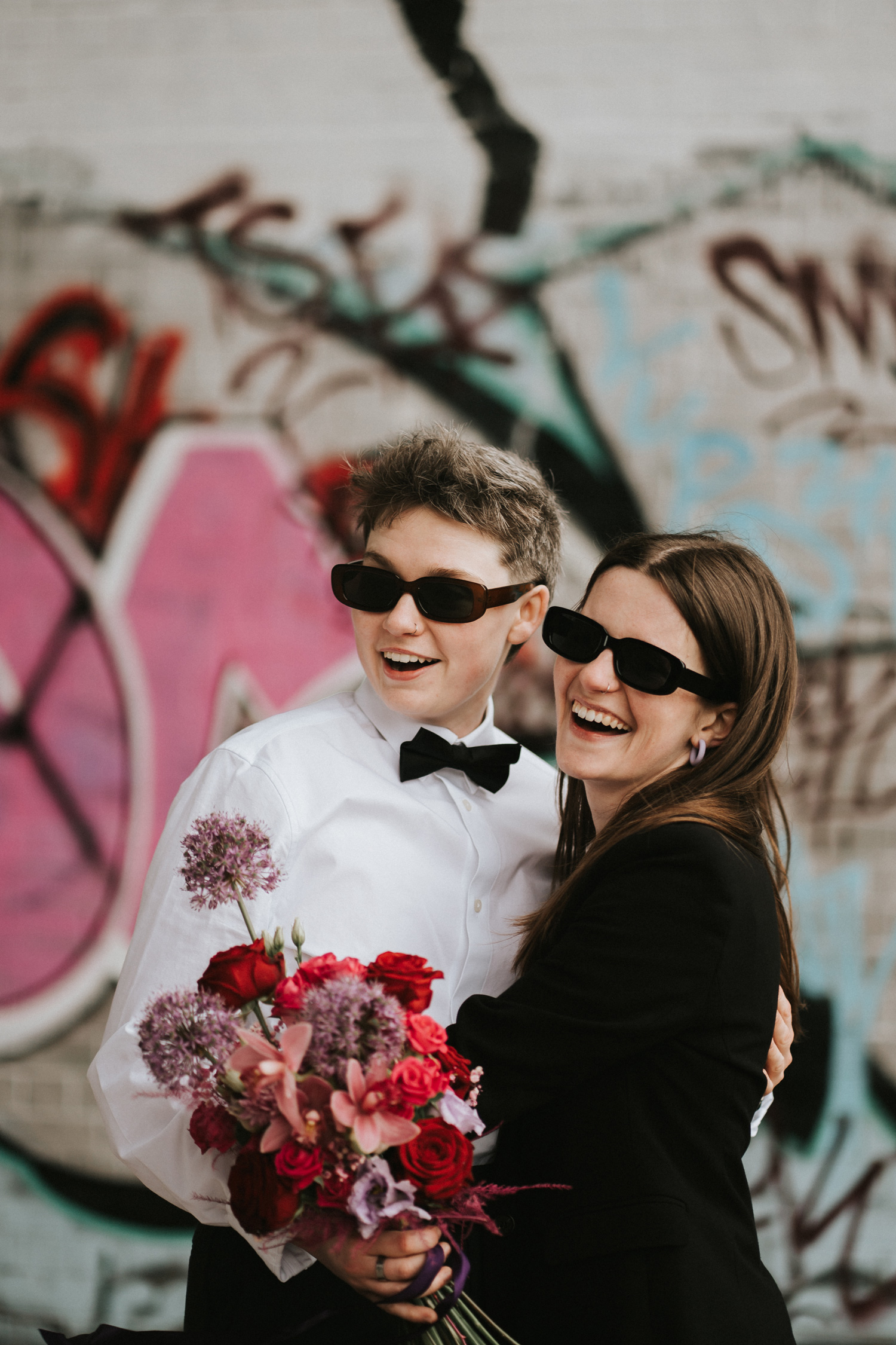 Newlyweds both wearing matching black sunglasses, smiling and holding a red bouquet in front of a graffiti wall. Shot by Steph Large