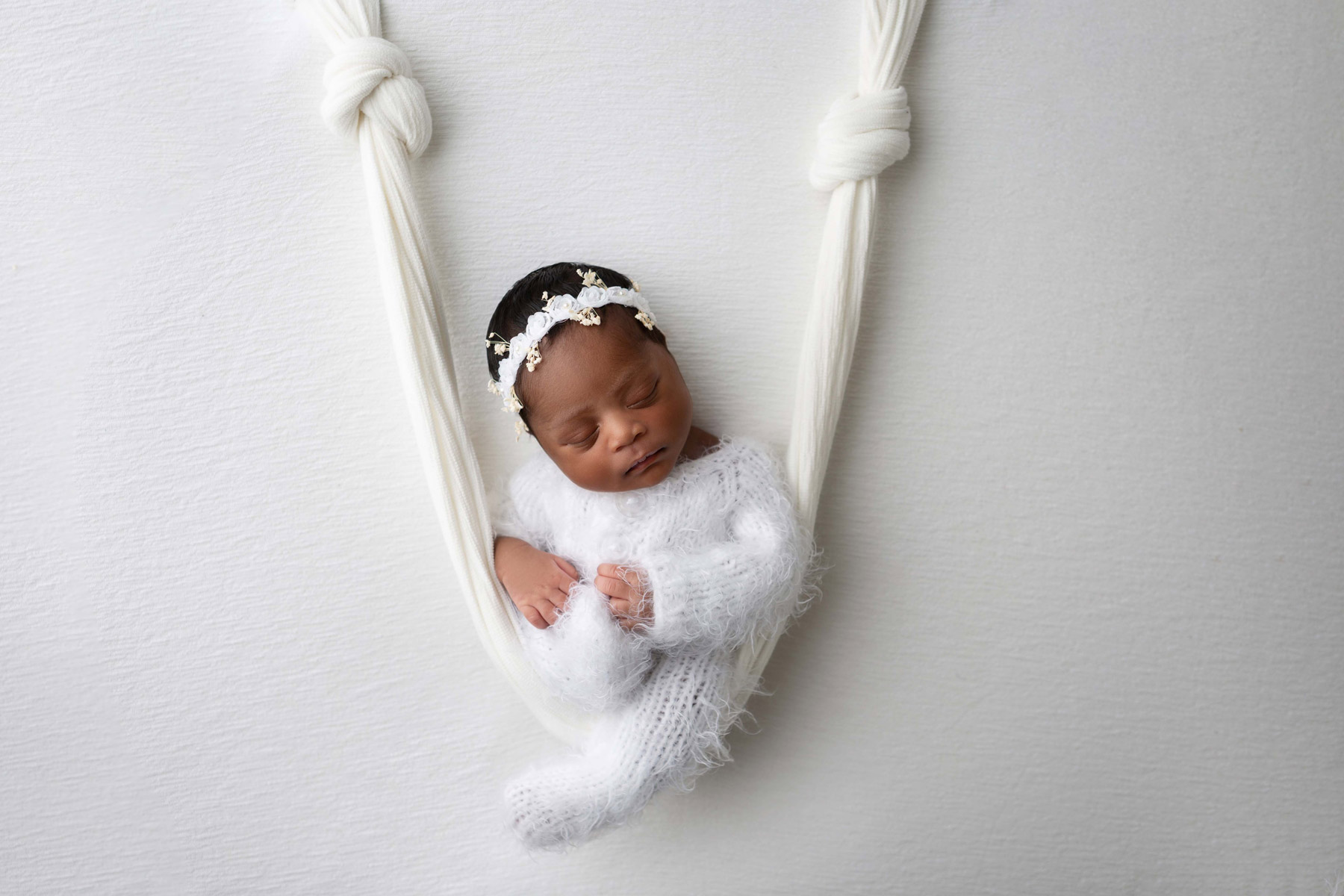 A maternity photo of a baby wearing a white headband who looks like they’re balancing on a white swing against a white background, shot by Jamella Binns with a Canon EOS 5D Mark III