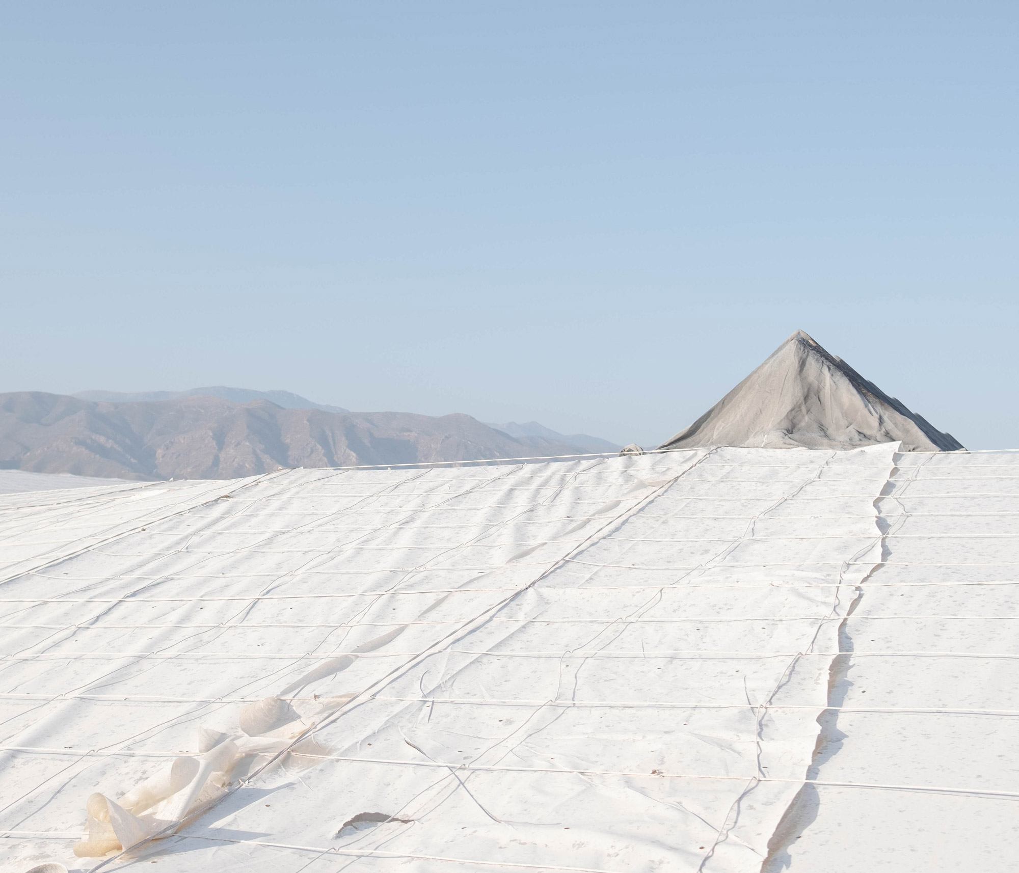 A mountain peak rises above a landscape of artificial white sheet material with a blue sky overhead, shot by Jordi Jon Pardo with a Fujifilm X-Pro 2