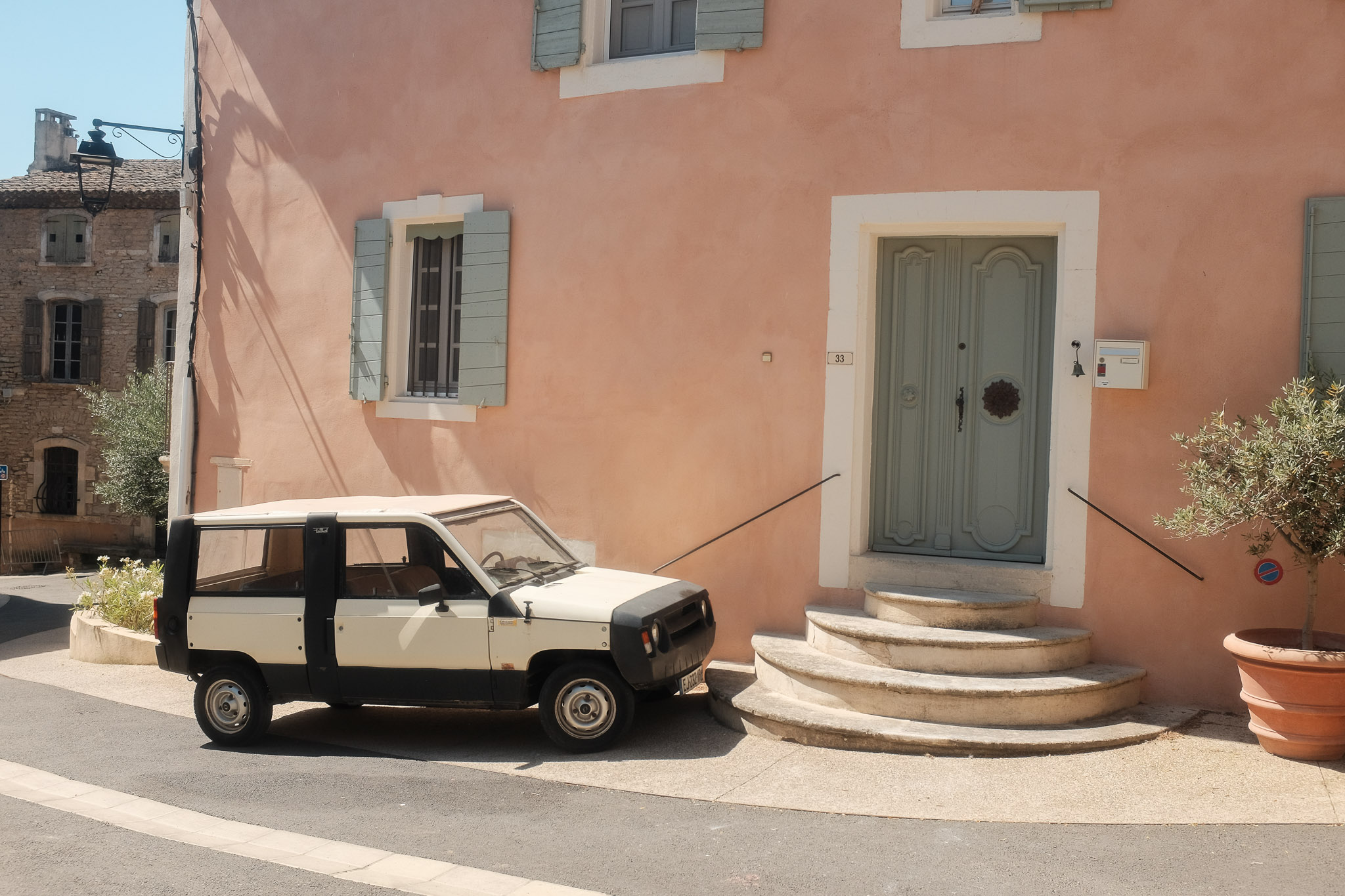 A small vintage boxy car is parked by a light pink and baby blue house in Gordes, France. Taken on the Fujifilm X100T by Connor Redmond.