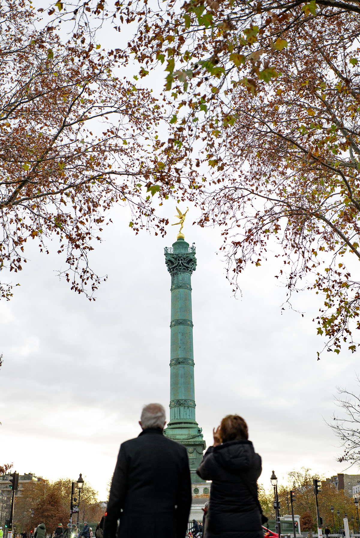 The July Column at Place de la Bastille in Paris framed by autumn trees, with people in the foreground under a soft overcast sky.