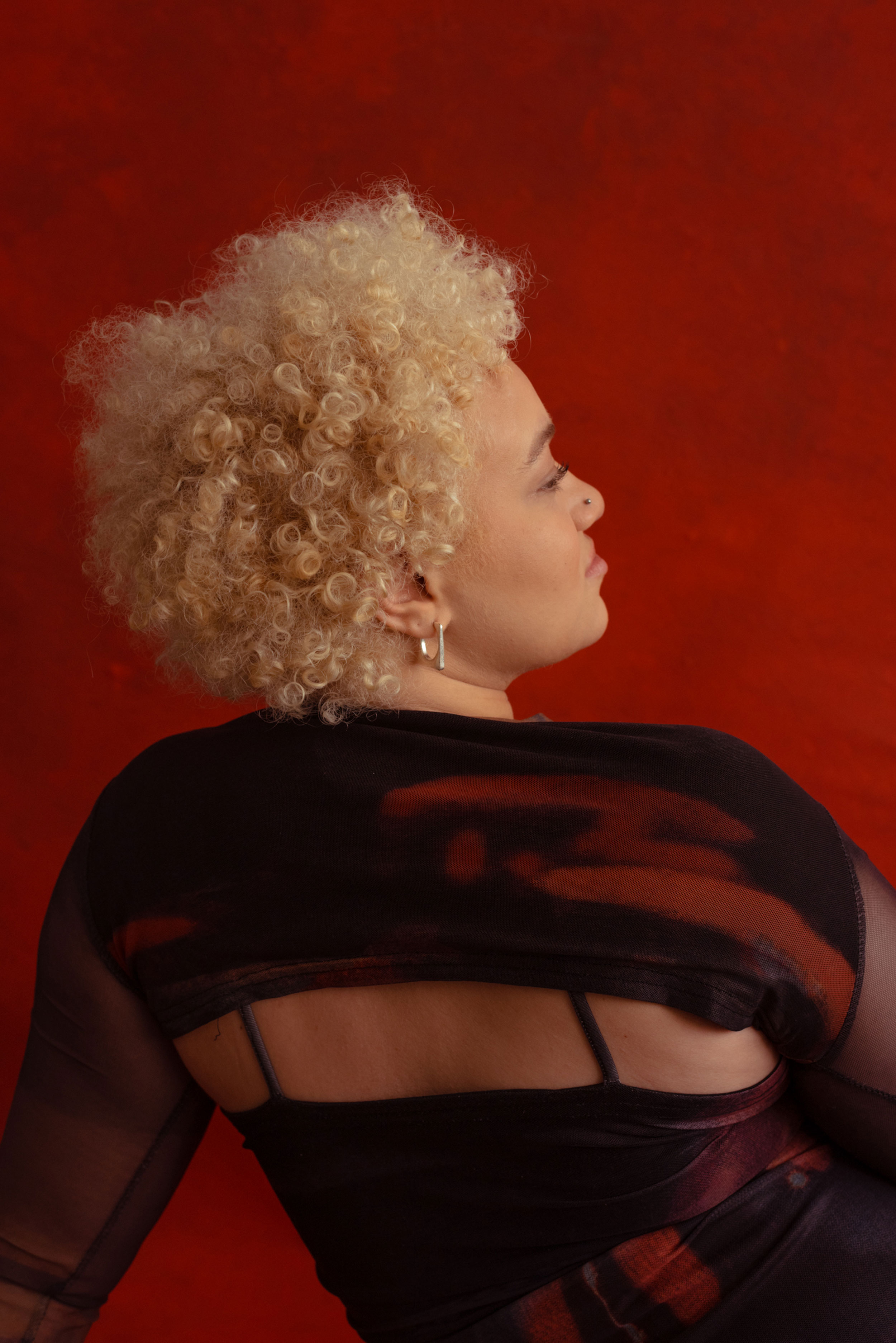 Woman with blond afro photographed from behind, looking to the right in front of a red background, shot by Nicole Benewaah