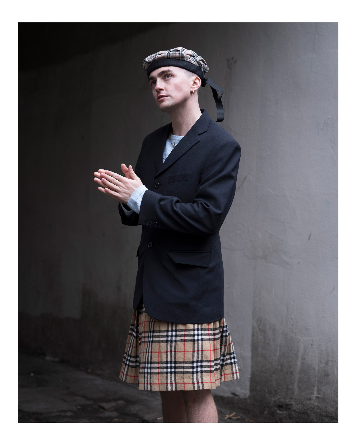 Farbporträt eines Mannes, der einen Kilt, einen Tartan Tam o' Shanter-Hut und einen Blazer trägt, seine Hände flach zusammenhält und vor einer grauen Wand steht, fotografiert von Simon Murphy mit einer Fujifilm GFX 100.