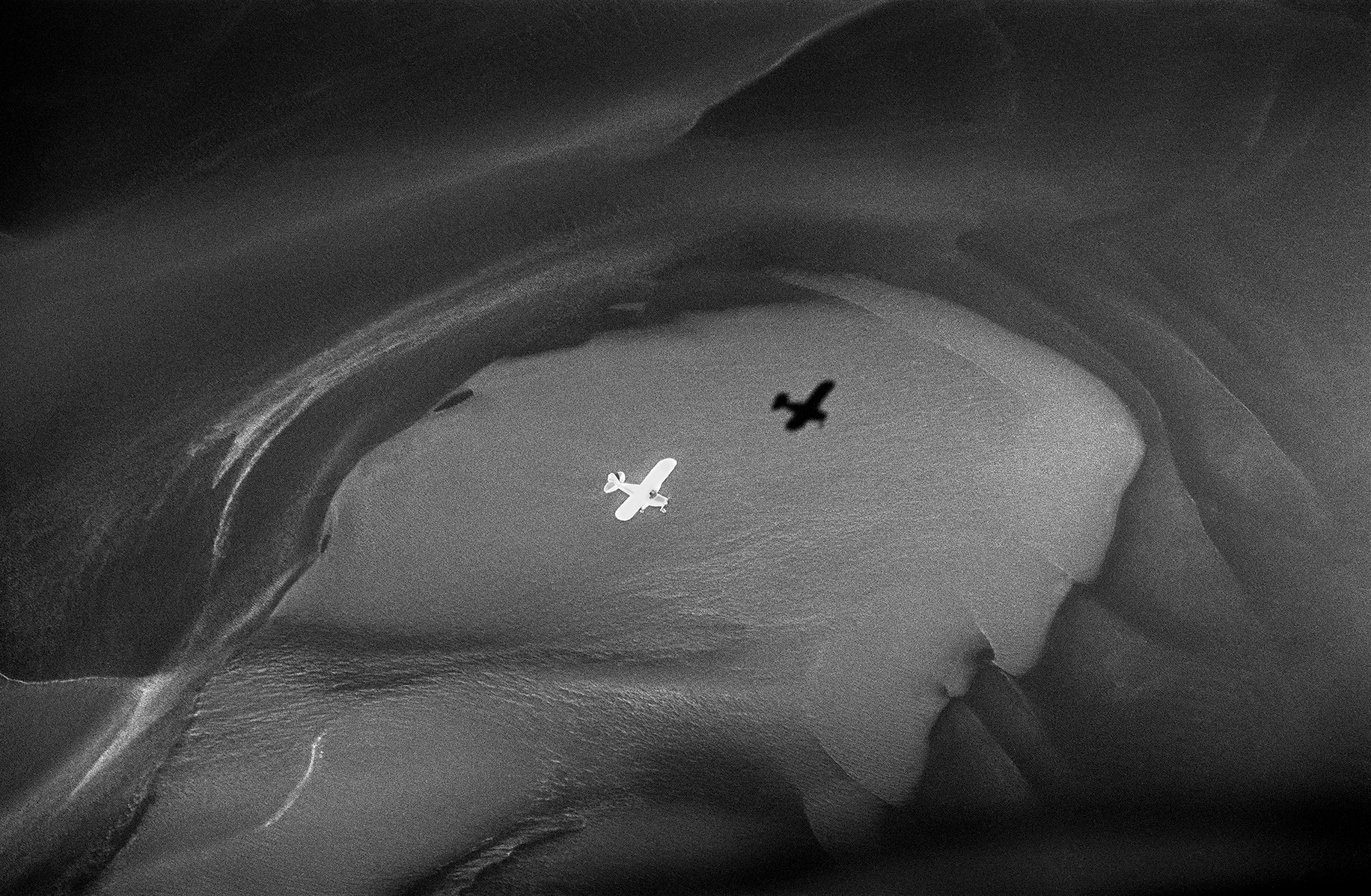 Black and white aerial view of a small plane from above, the plane reflected in a small lake below, photo taken by Ragnar Axelsson