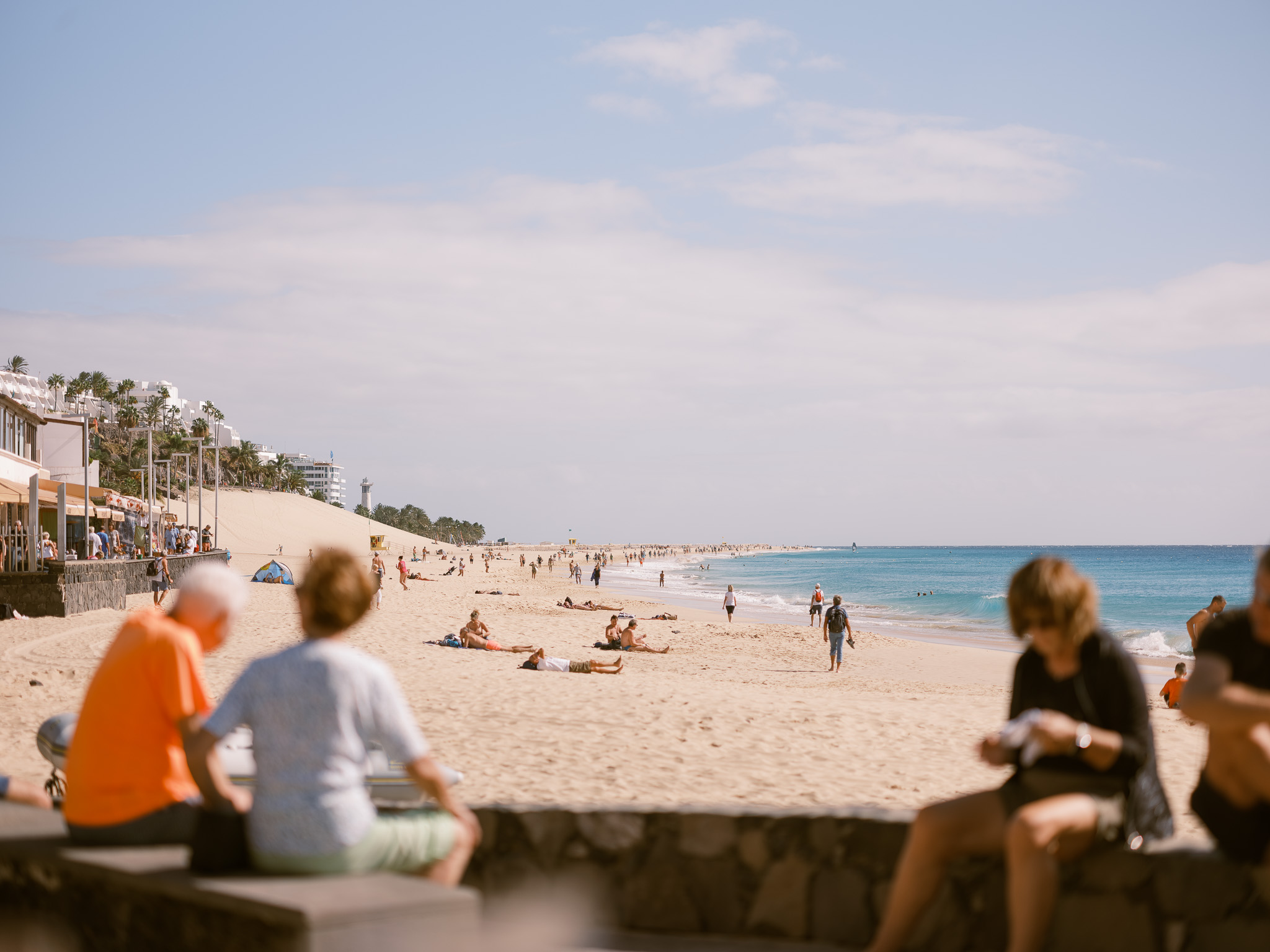Ein Strand mit sonnenbadenden und spazierenden Menschen, mit unscharfen Menschen im Vordergrund von Connor Redmond

