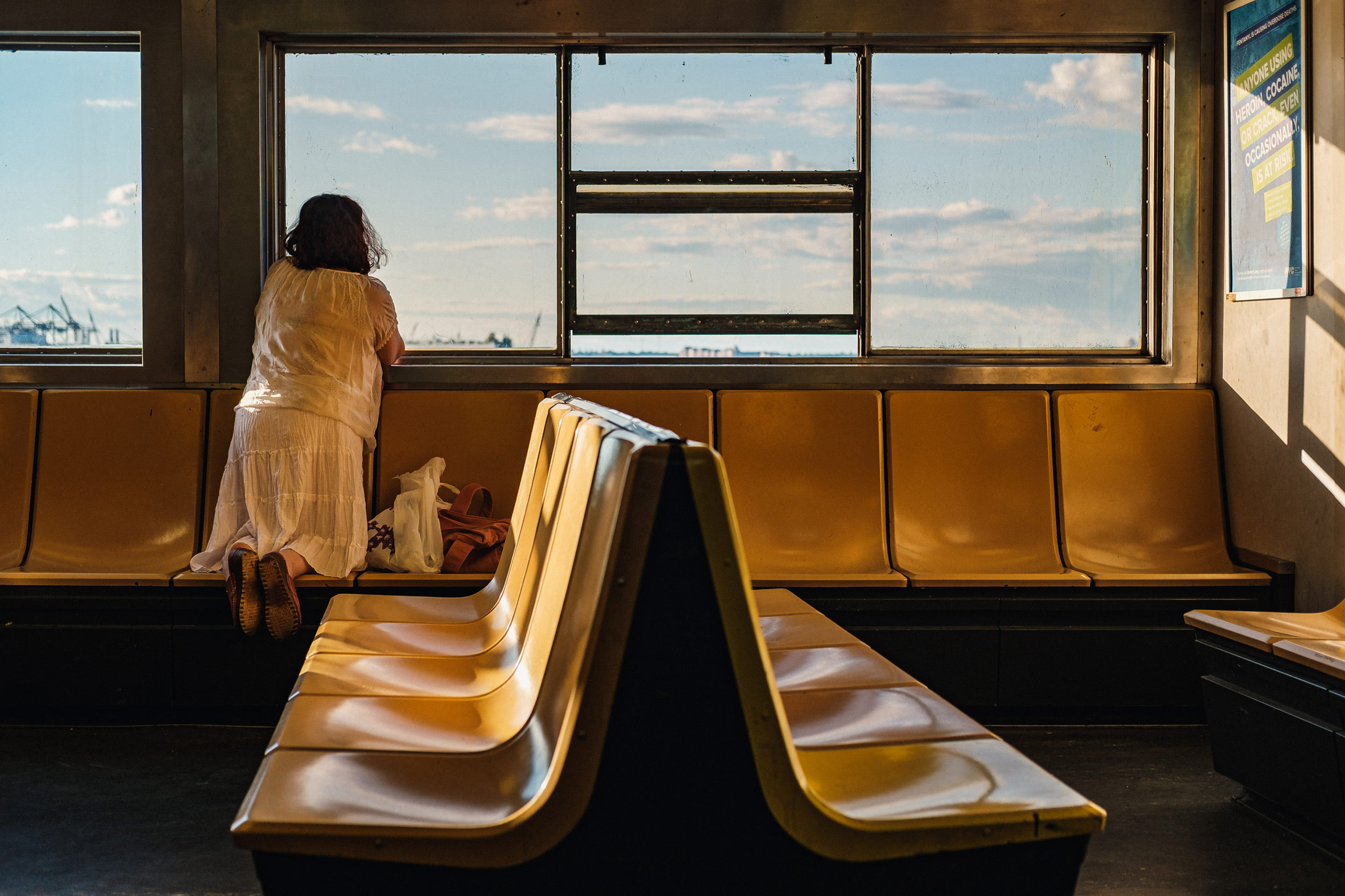 Persoon leunt tegen stoel en kijkt uit raam van Staten Island Ferry in NYC met blauwe lucht op achtergrond, gefotografeerd door Manuel Pena.