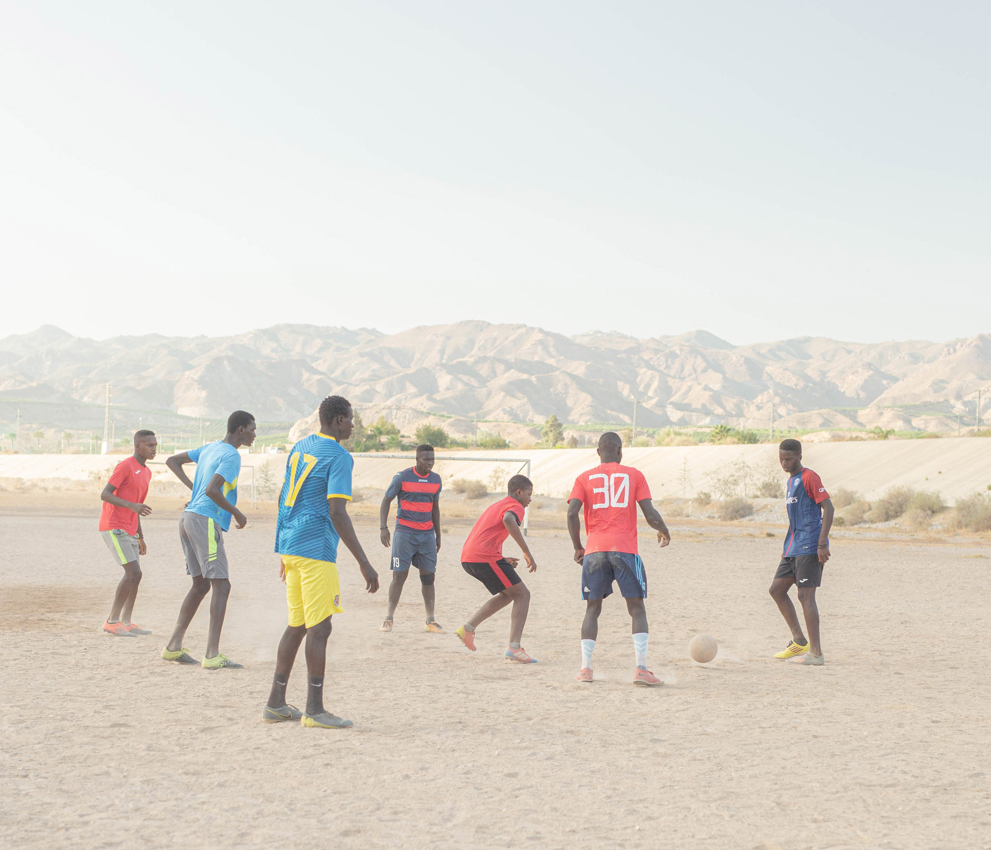 Seven people wearing different football kits playing football on sand surrounded by mountains and a pale blue sky, shot by Jordi Jon Pardo with a Canon EOS 5D Mark III