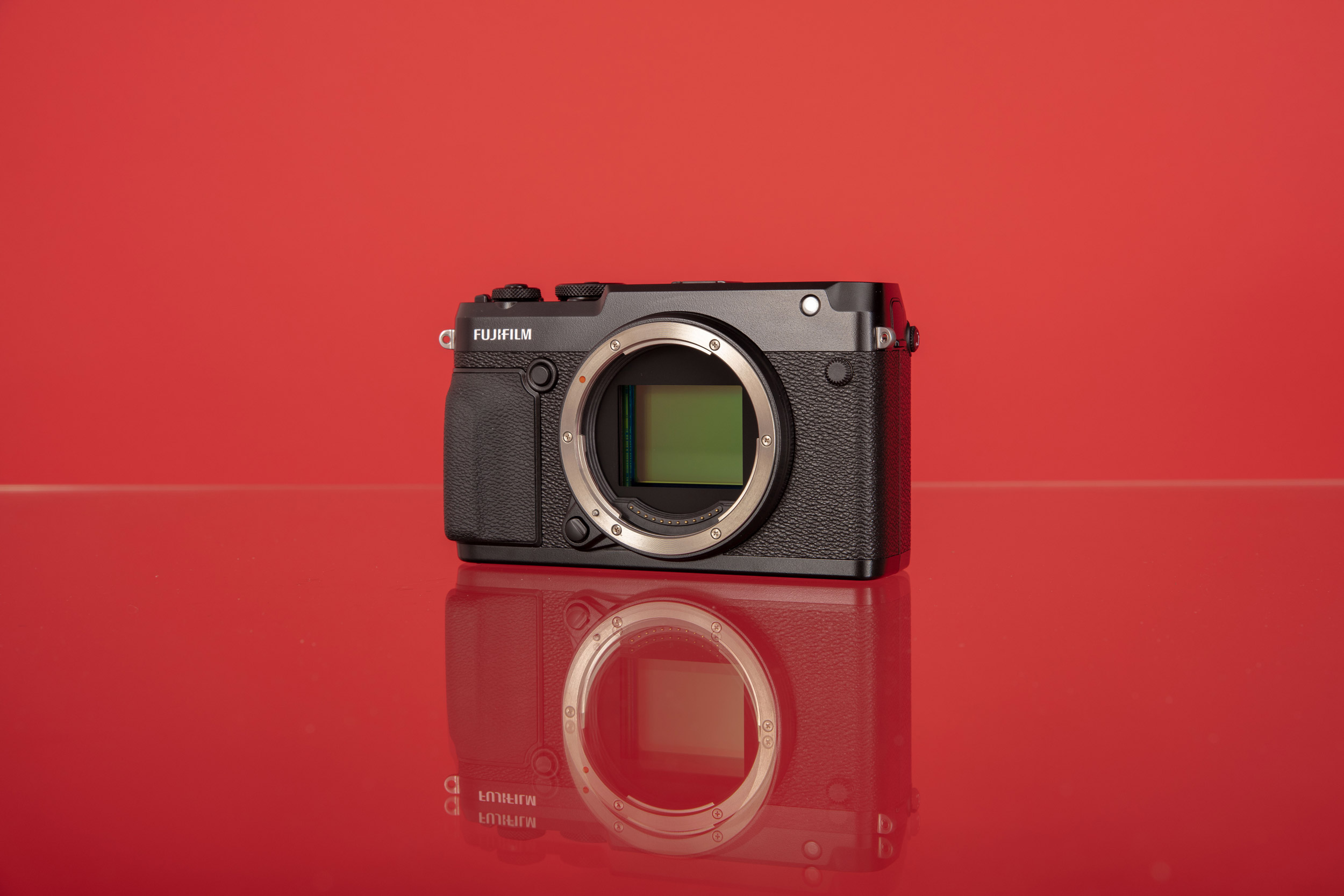 A photo of a Fujifilm GFX 50R medium-format digital camera with its sensor exposed against a red background reflecting onto a glass table