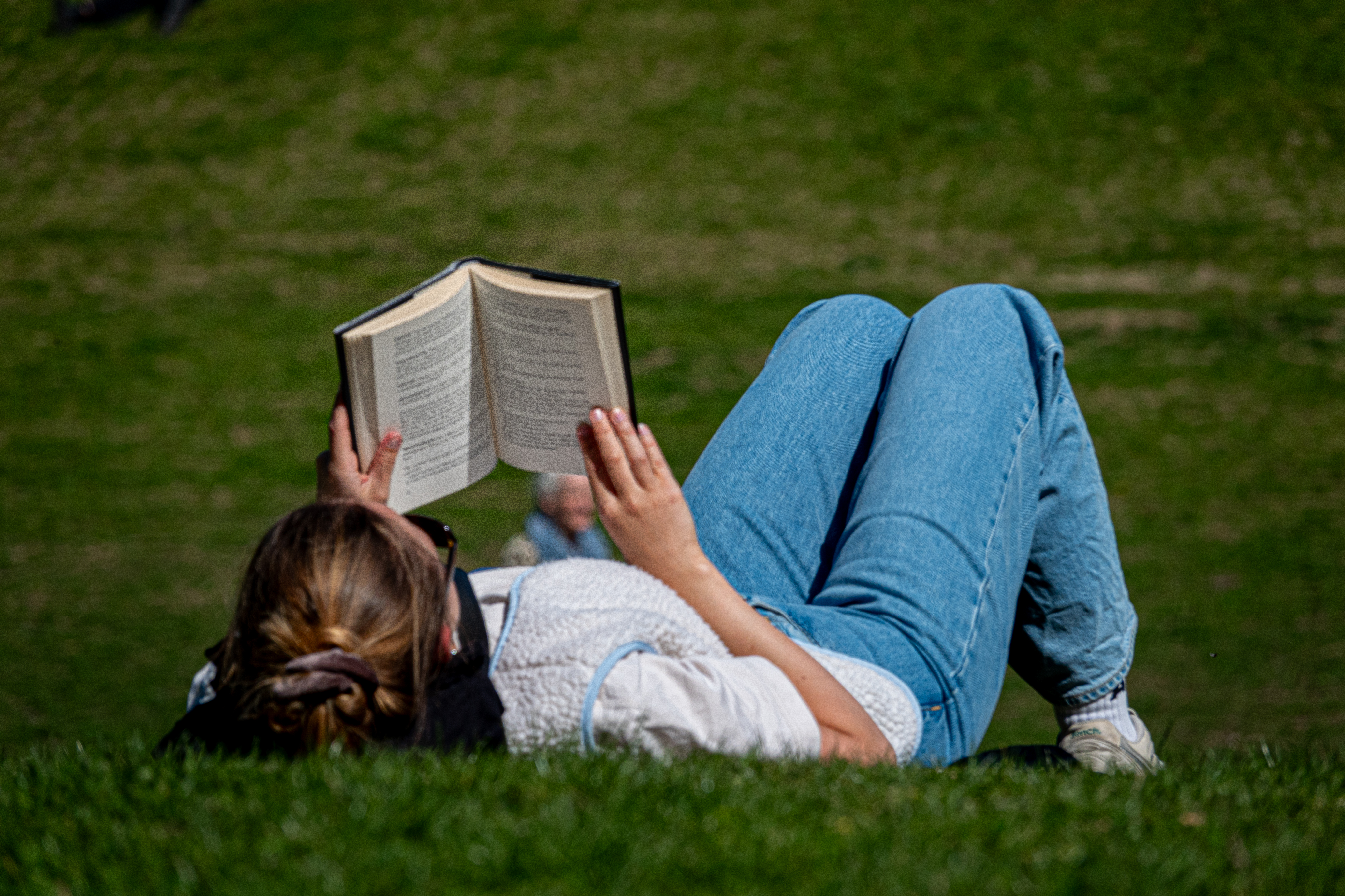 Una chica lee un libro tumbada en la hierba en un parque soleado.