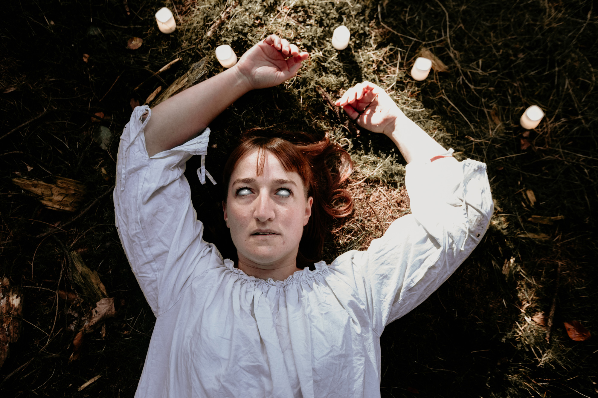 Horror colour photograph of a model in a white dress lying on the forest floor with pupils removed in post-production 

