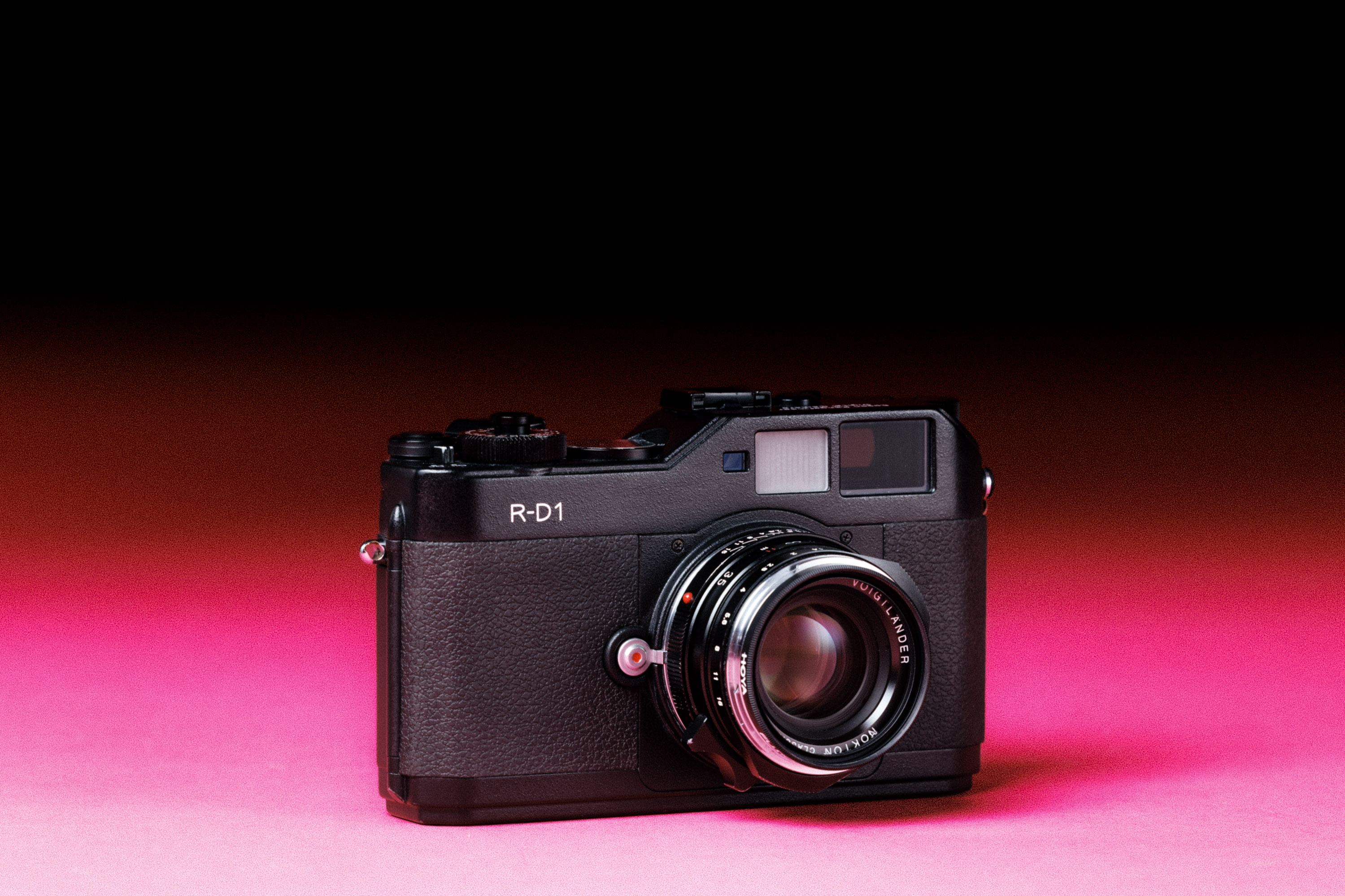 A used Epson R-D1 camera with a Voigtlander Nokton Classic MC 35mm f/1.4 II VM lens, with an added film-like grain effect on a graduated pink background.