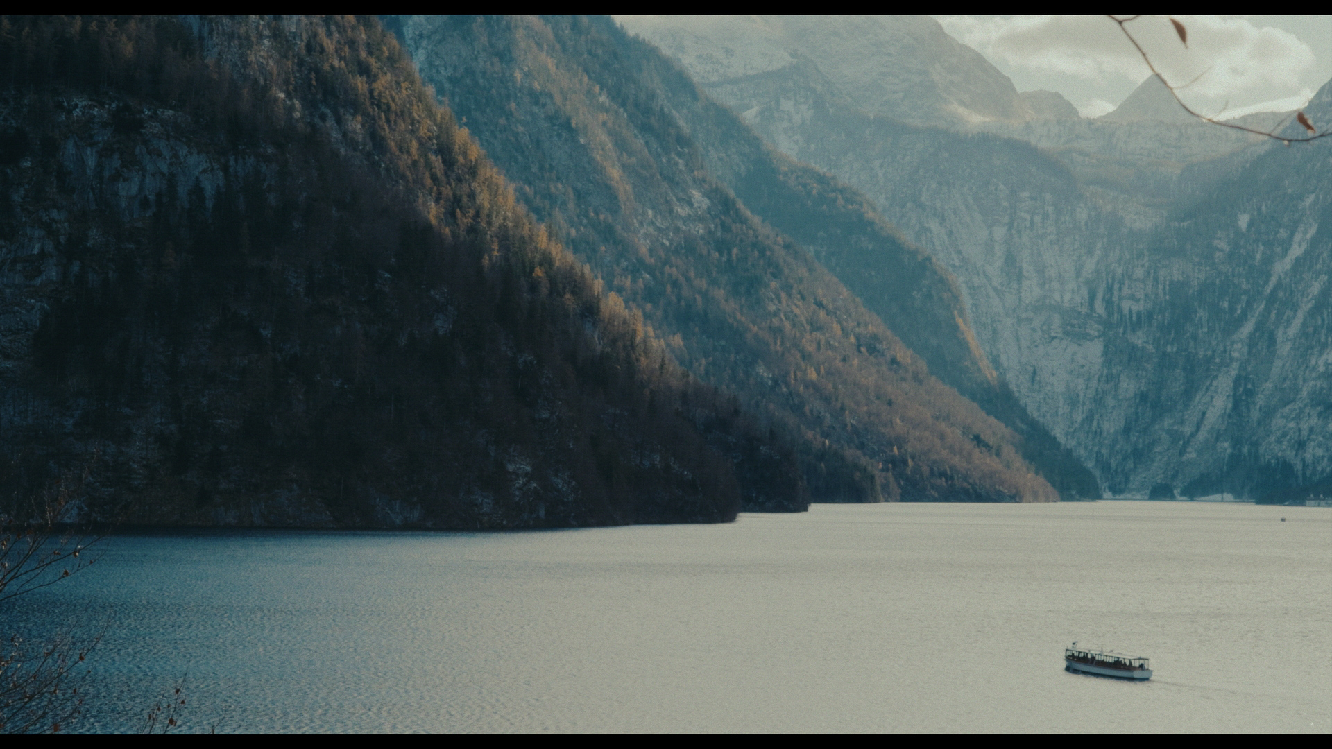 A small passenger boat navigates a vast lake surrounded by steep, dark, and snow-dusted mountain slopes under an overcast sky.