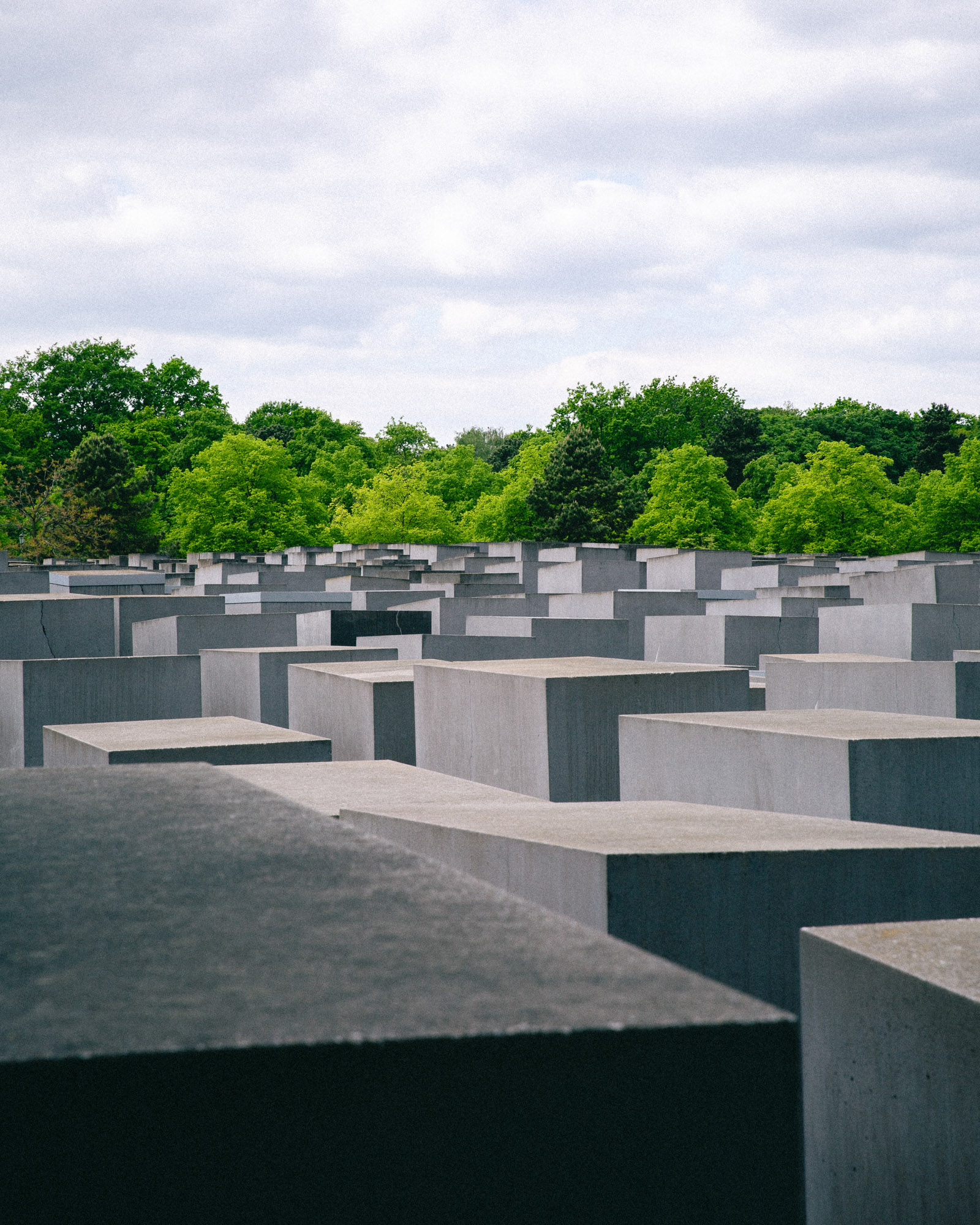 Photograph of Berlin’s Holocaust Memorial, with rows of concrete stelae stretching toward green trees beneath a cloudy sky. Shot by Austin Fassino on a Canon EOS 5D Mark III.