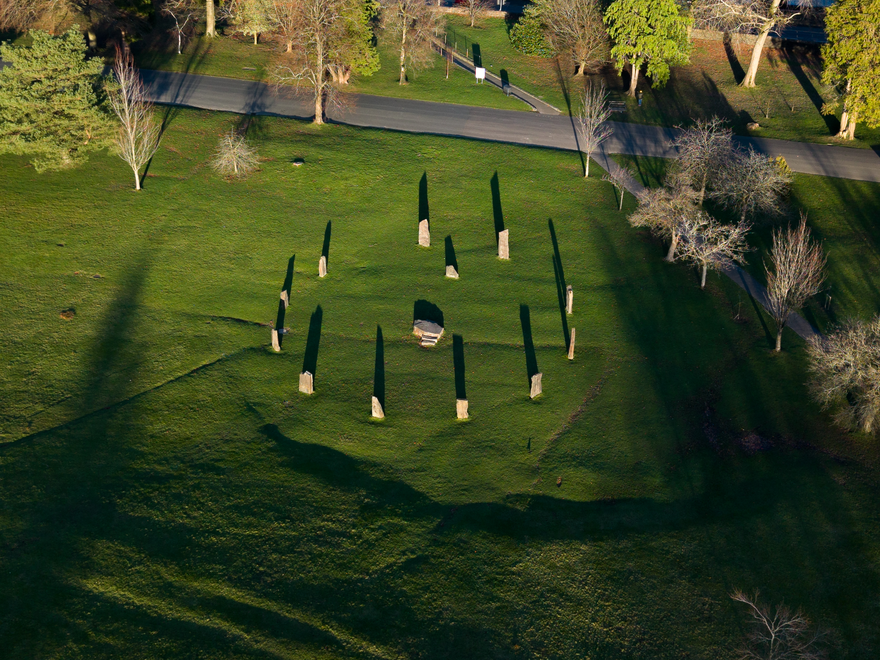 Imagem aérea captada com o DJI Mini 5 Pro num parque verde com árvores, caminhos e uma instalação circular de pedras verticais. A luz baixa cria sombras longas sobre a relva, dando profundidade e textura à composição.