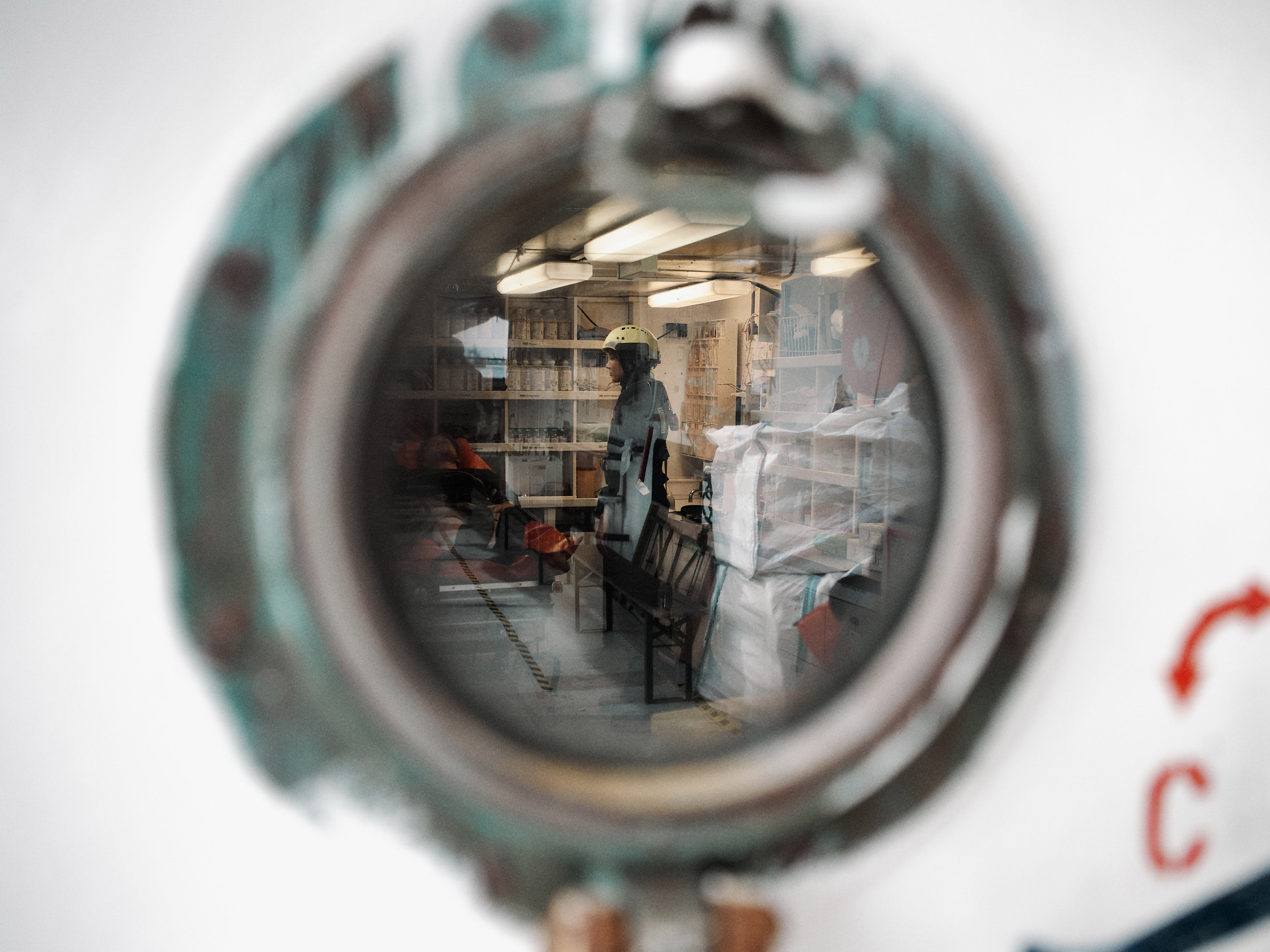 Photo through the porthole of a ship, behind a person wearing a helmet in a storage room.