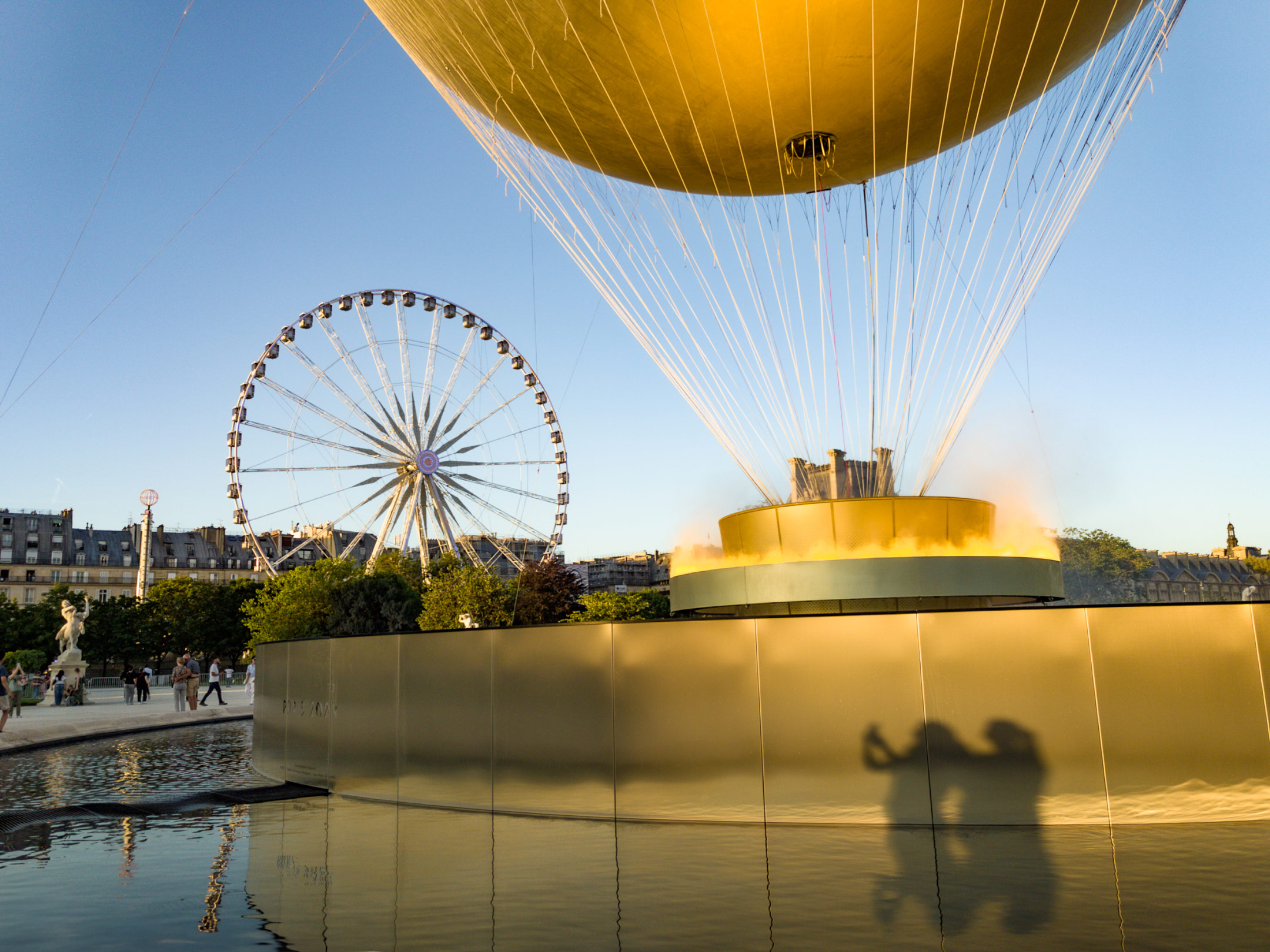 Golden tethered balloon in Jardin des Tuileries with a Ferris wheel in the background, reflecting on water during warm evening light in Paris.