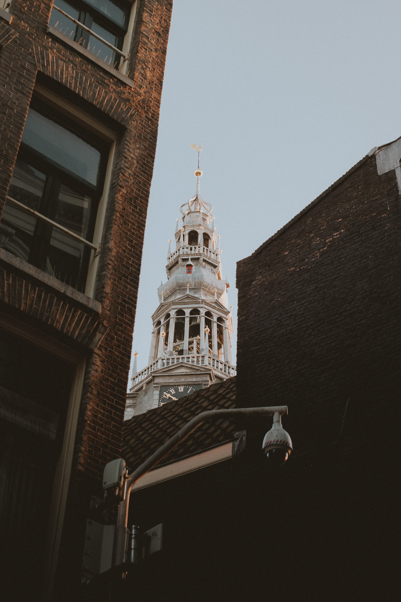 View of an Amsterdam church tower framed by narrow brick buildings, captured from a low angle with warm light highlighting classic Dutch architecture.