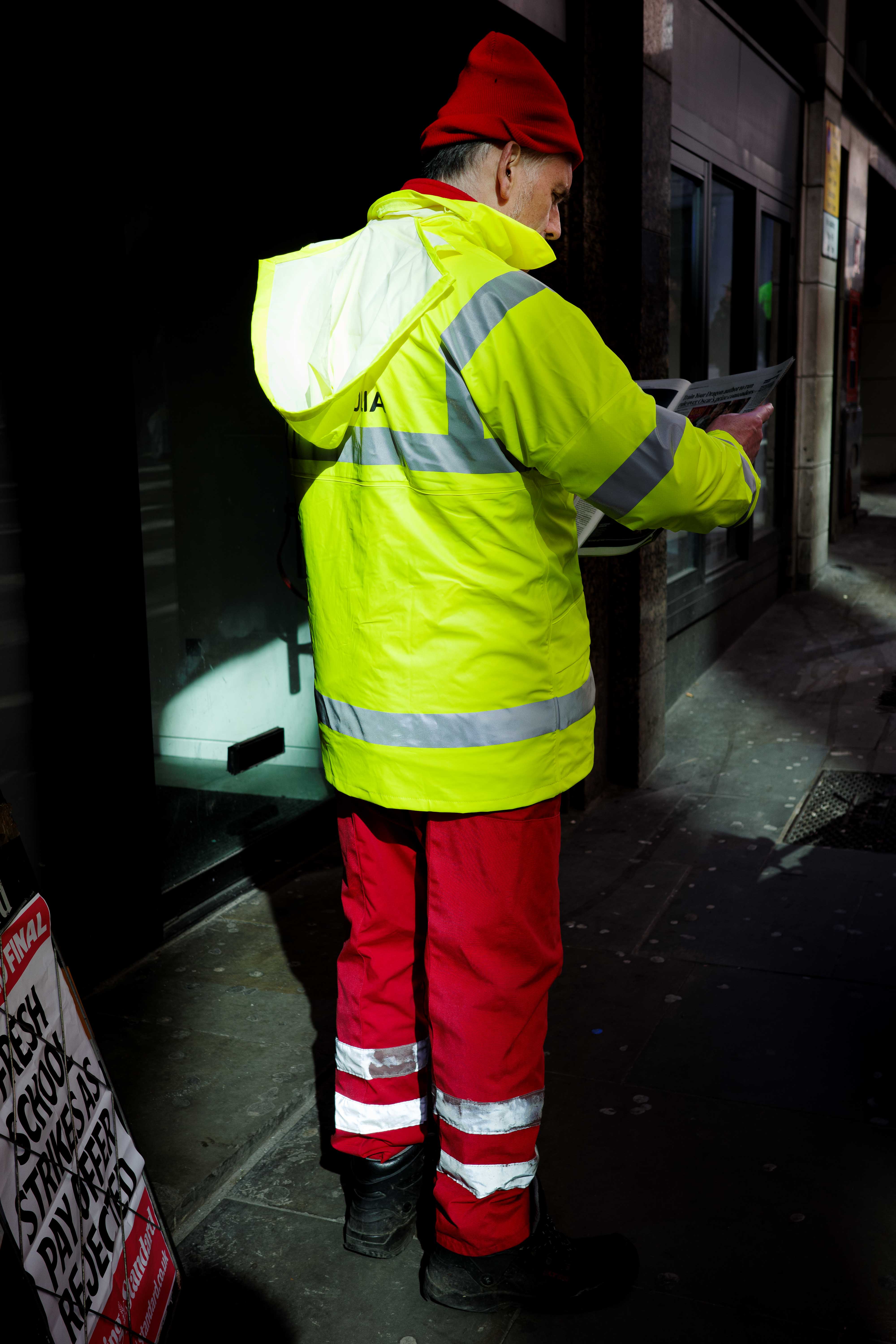Person with a high-vis jacket, red trousers and a red hat reads a newspaper on the street